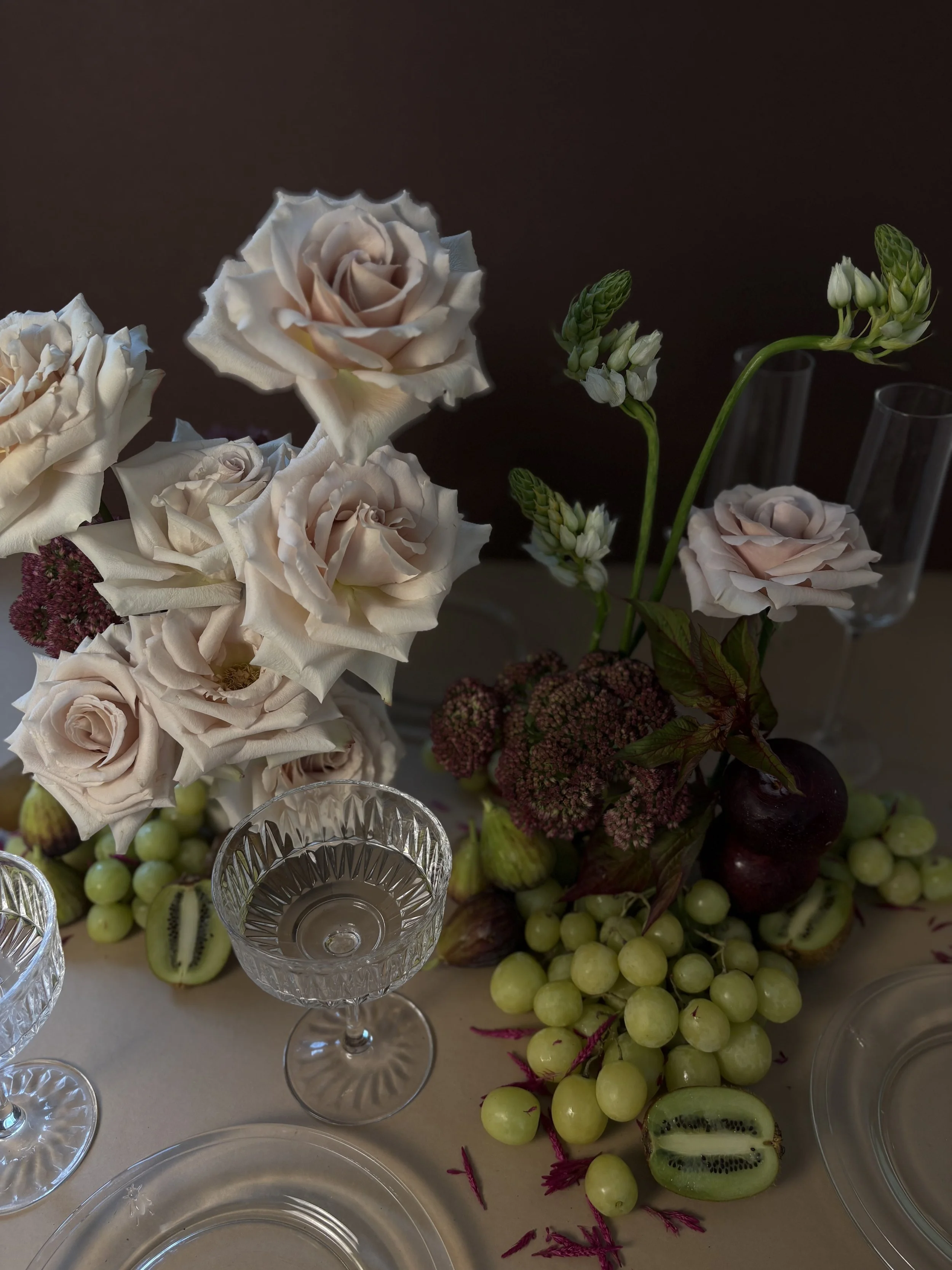 Elegant floral centerpiece with cream-colored roses and white lilies, surrounded by assorted fruits including grapes, kiwi, and an apple, on a table with glassware and plates.