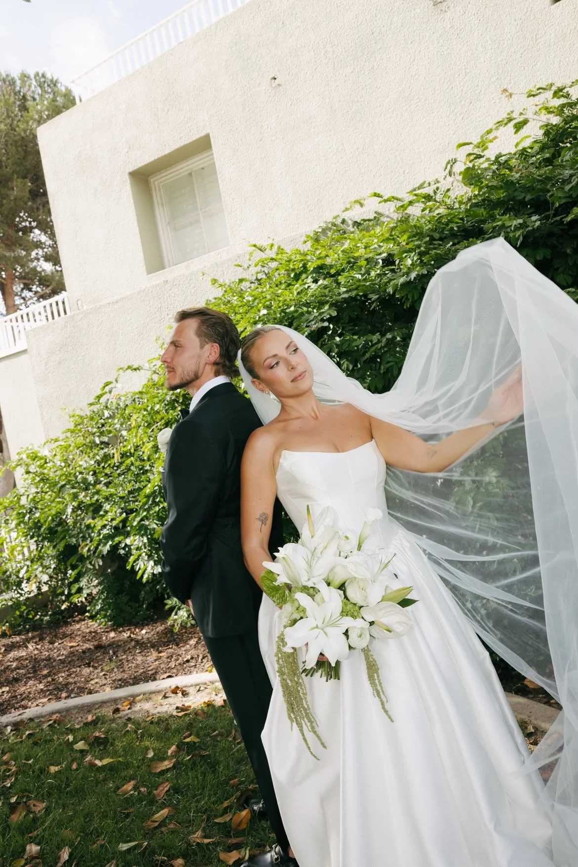 A bride in a white wedding dress holding a bouquet of white lilies and greenery, standing next to a groom in a black suit outdoors with a white building and green bushes in the background.