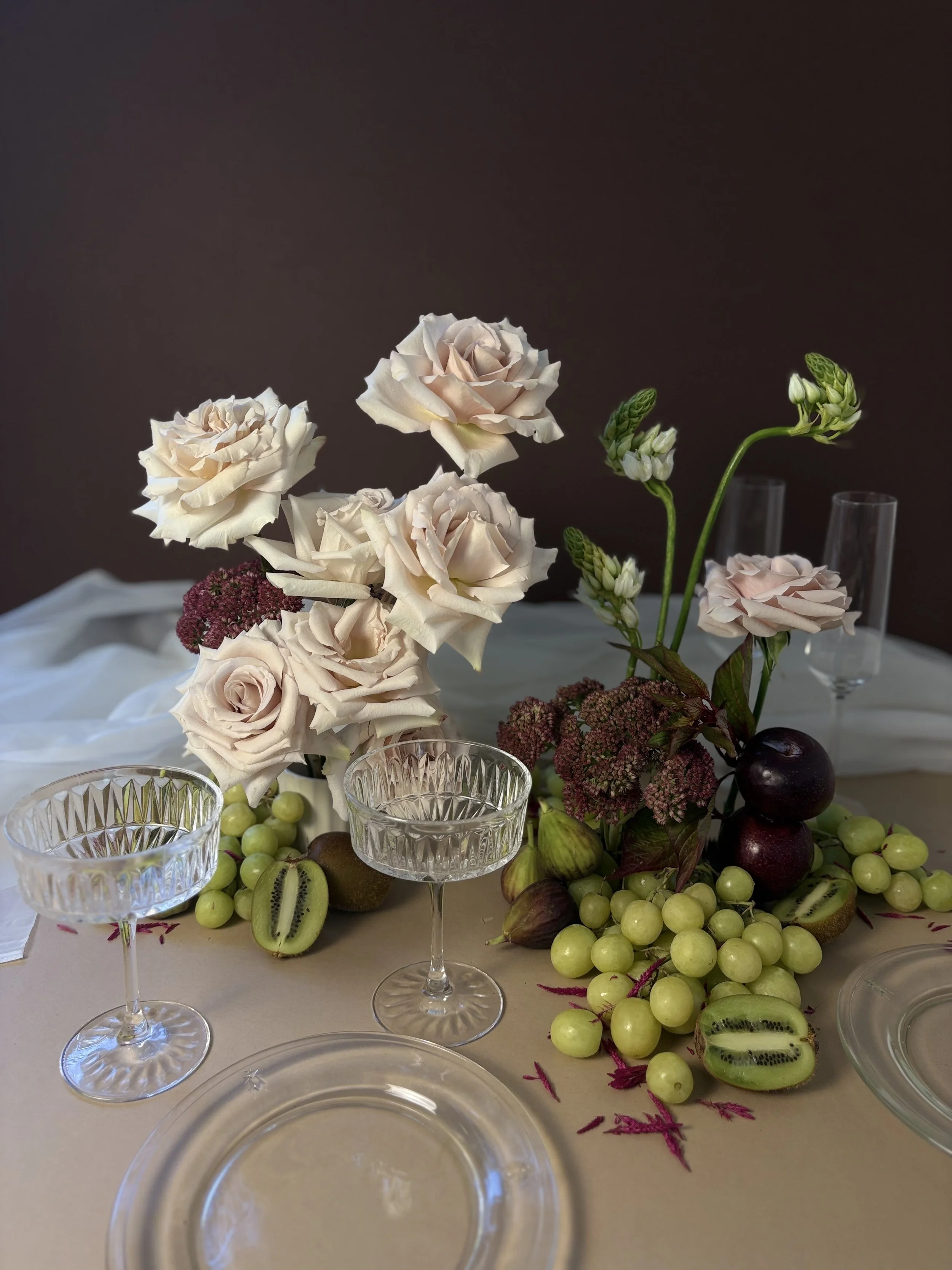 A table decorated with white and pink roses, grapes, figs, and empty glassware against a dark background.
