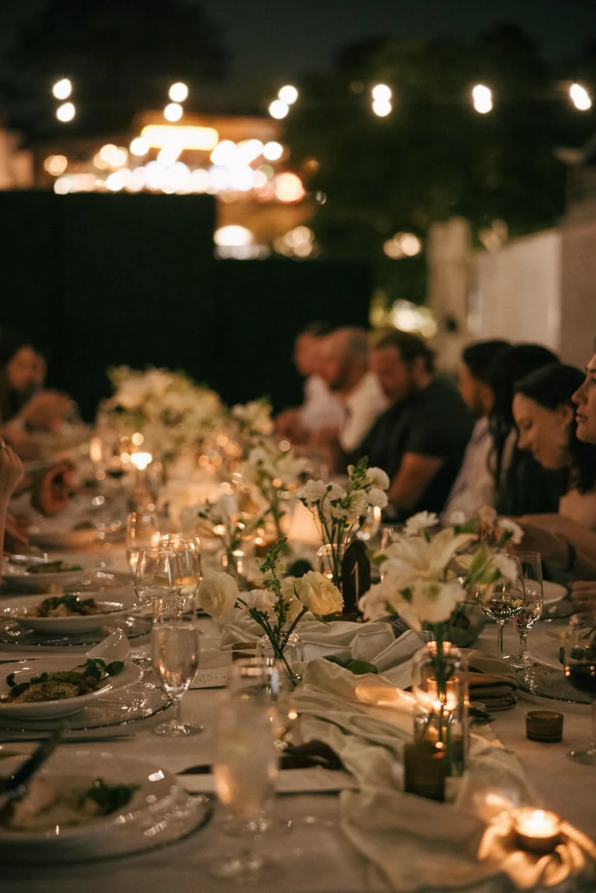 A nighttime dinner event featuring a long elegantly set table decorated with white flowers and lit candles, with guests seated and engaged in conversation.