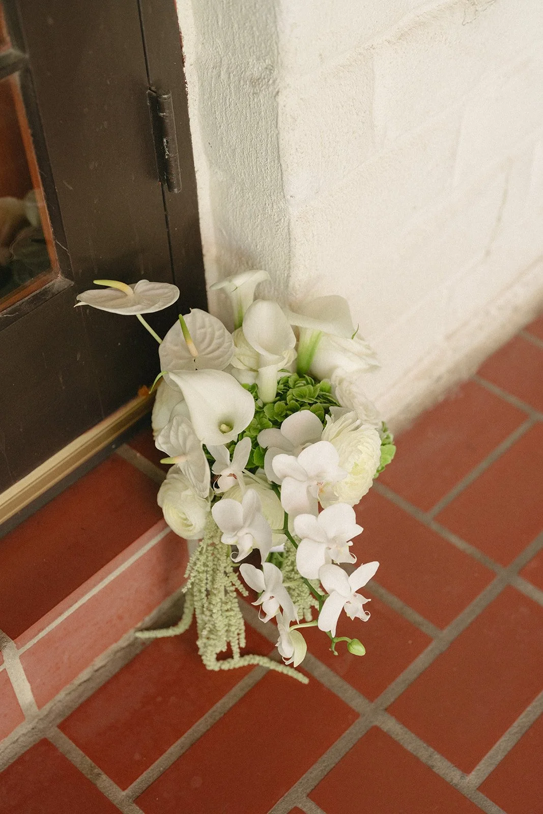 A bouquet of white flowers including orchids and calla lilies, placed on a tiled floor next to a dark-colored door and a white wall.