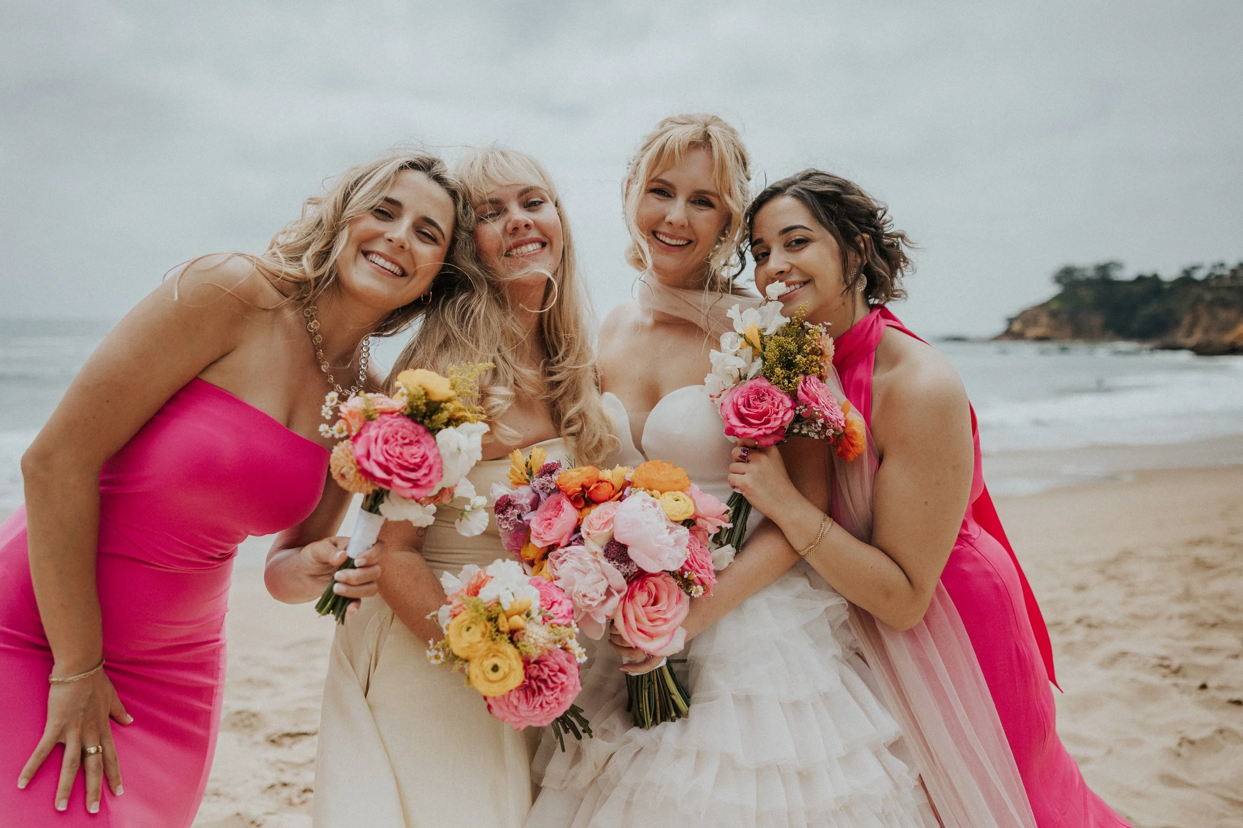 Four women posing together on a beach, with three holding bouquets of flowers. The woman in the center is wearing a white wedding dress, indicating she is the bride, while the others are in colorful dresses, likely bridesmaids or friends.