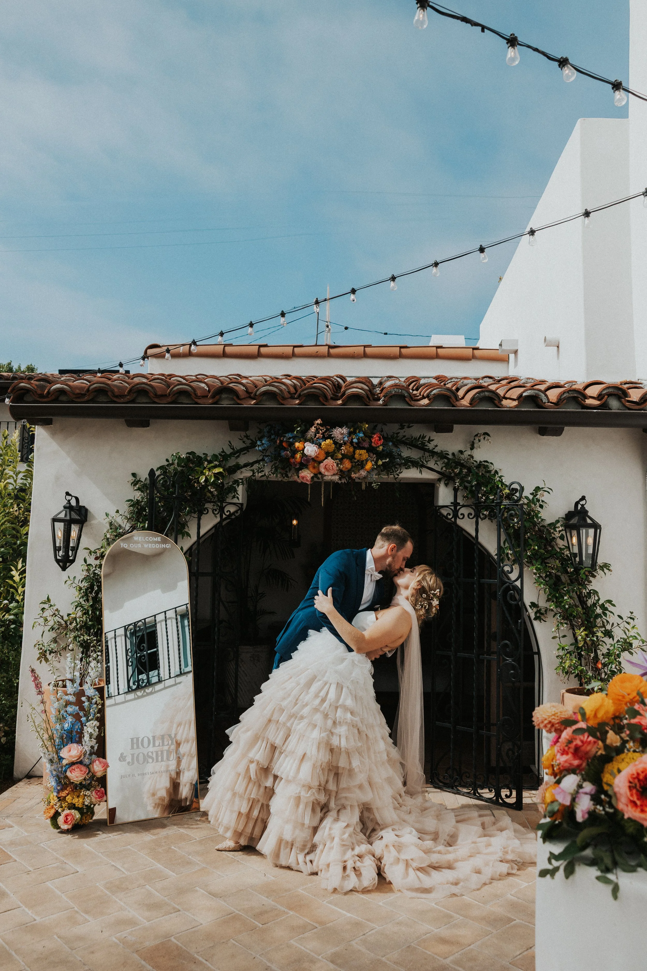 A bride and groom sharing a kiss outside a wedding venue decorated with flowers and outdoor lights.