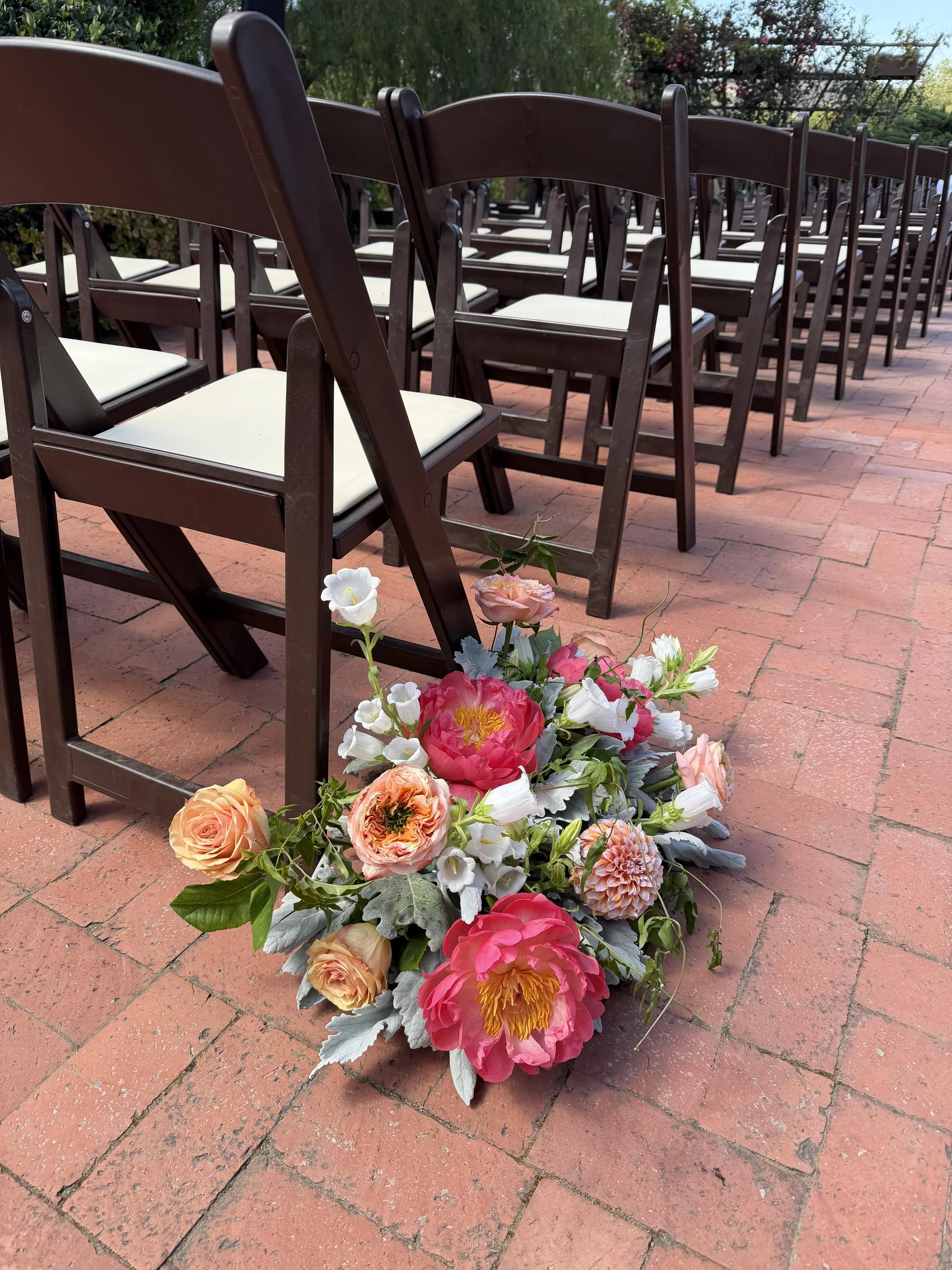 A line of brown wooden chairs with white cushions arranged on a brick patio. In the foreground, there is a bouquet of pink, white, and orange flowers with green leaves and some silvery foliage.