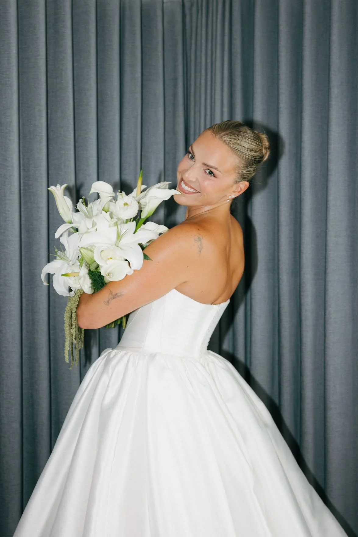 A woman in a strapless white wedding dress holding a bouquet of white lilies, smiling and standing in front of gray curtains.