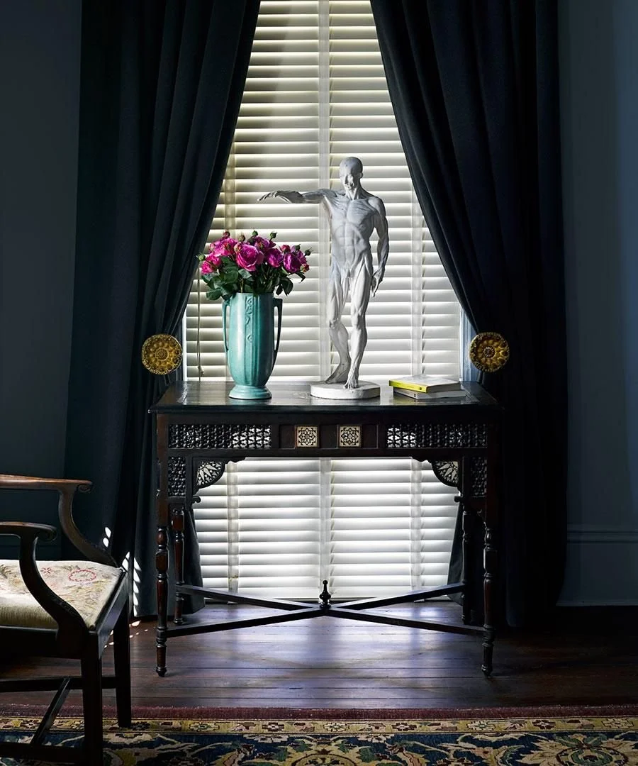 A window with white blinds and dark curtains partially open, displaying a decorative table beneath. On the table, there is a turquoise vase filled with pink roses, a white sculpture of a human figure extending one arm, a yellow book, and two small de