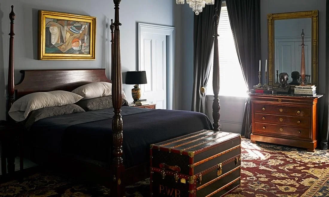 A bedroom with a four-poster bed, dark curtains, a chest at foot of bed, wooden dresser with mirror, and artwork on the wall.