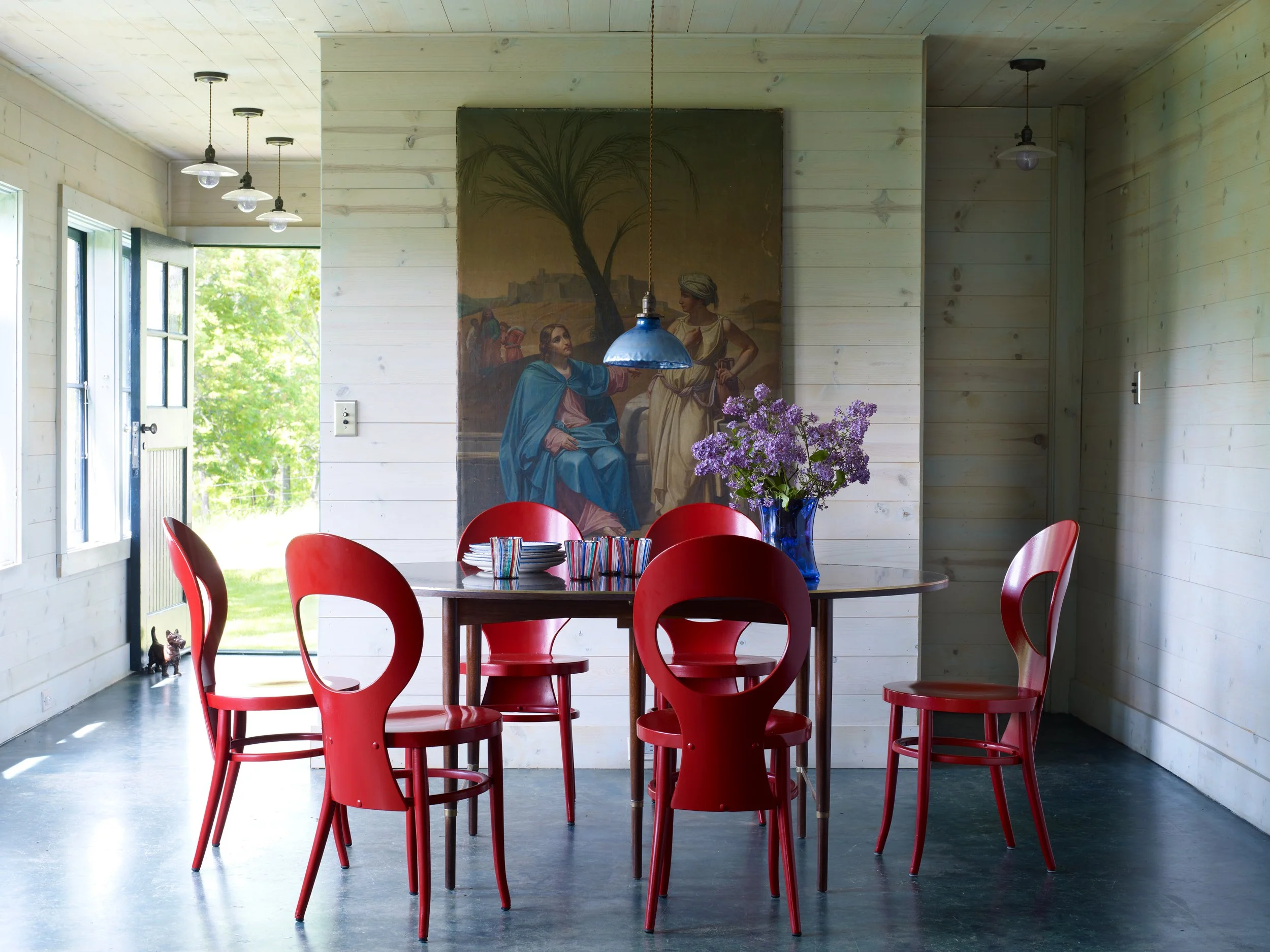 A dining room with a round wooden table surrounded by six red chairs. A blue vase with purple flowers is on the table. Behind the table, there is a large painting depicting a biblical scene with a seated man in blue robes and a standing woman with a 