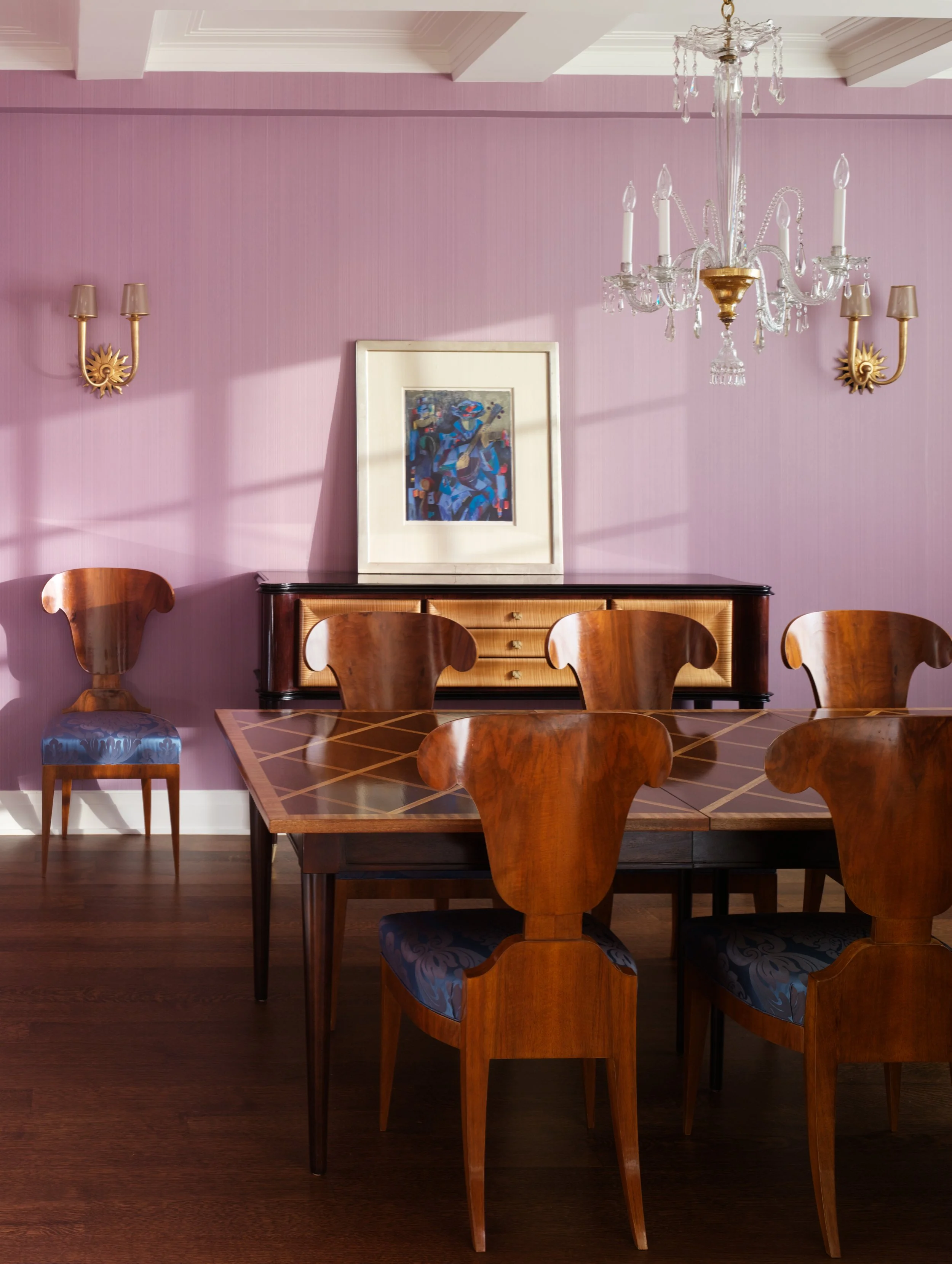 A dining room with pink walls, a chandelier, and wooden dining chairs surrounding a wooden table. There is a sideboard with a framed picture on it and wall-mounted light fixtures.