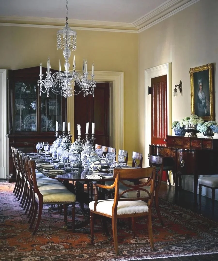 Elegant dining room with a long wooden table set with glassware and candlesticks, chandelier hanging above, wooden cabinet in corner, portrait on wall, and decorative floral arrangements.