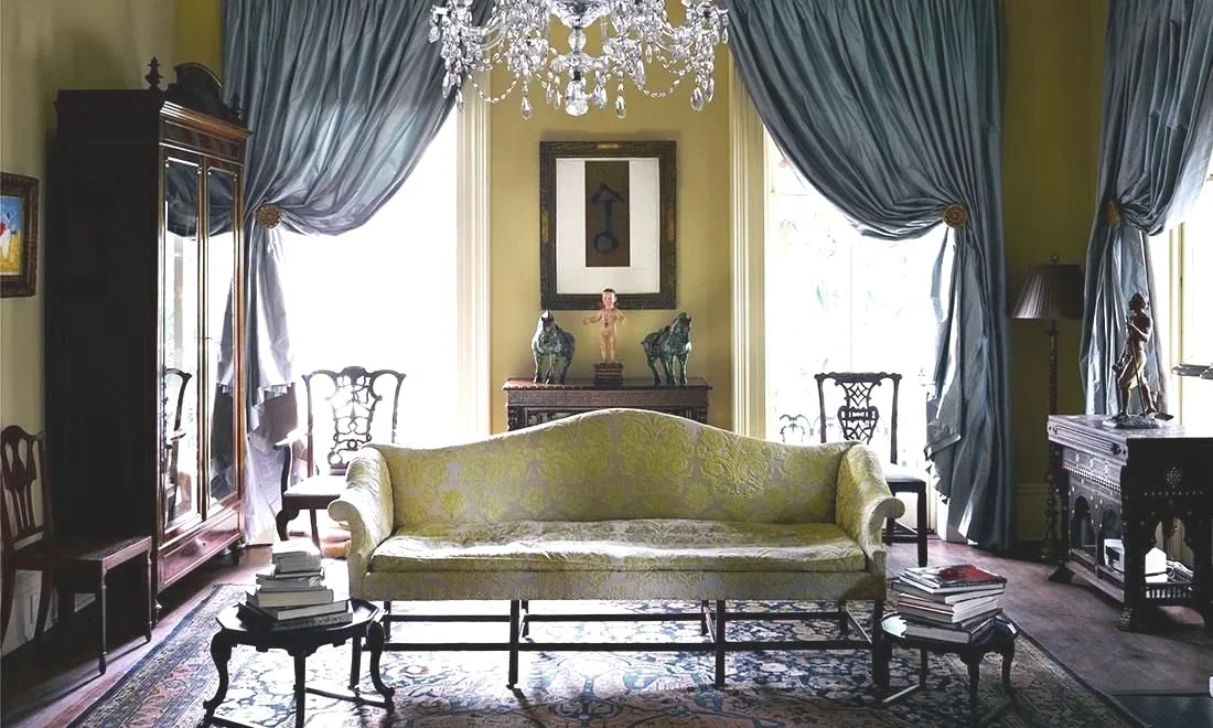 Elegant living room with large windows, blue drapes, a chandelier, antique furniture, and decorative sculptures.