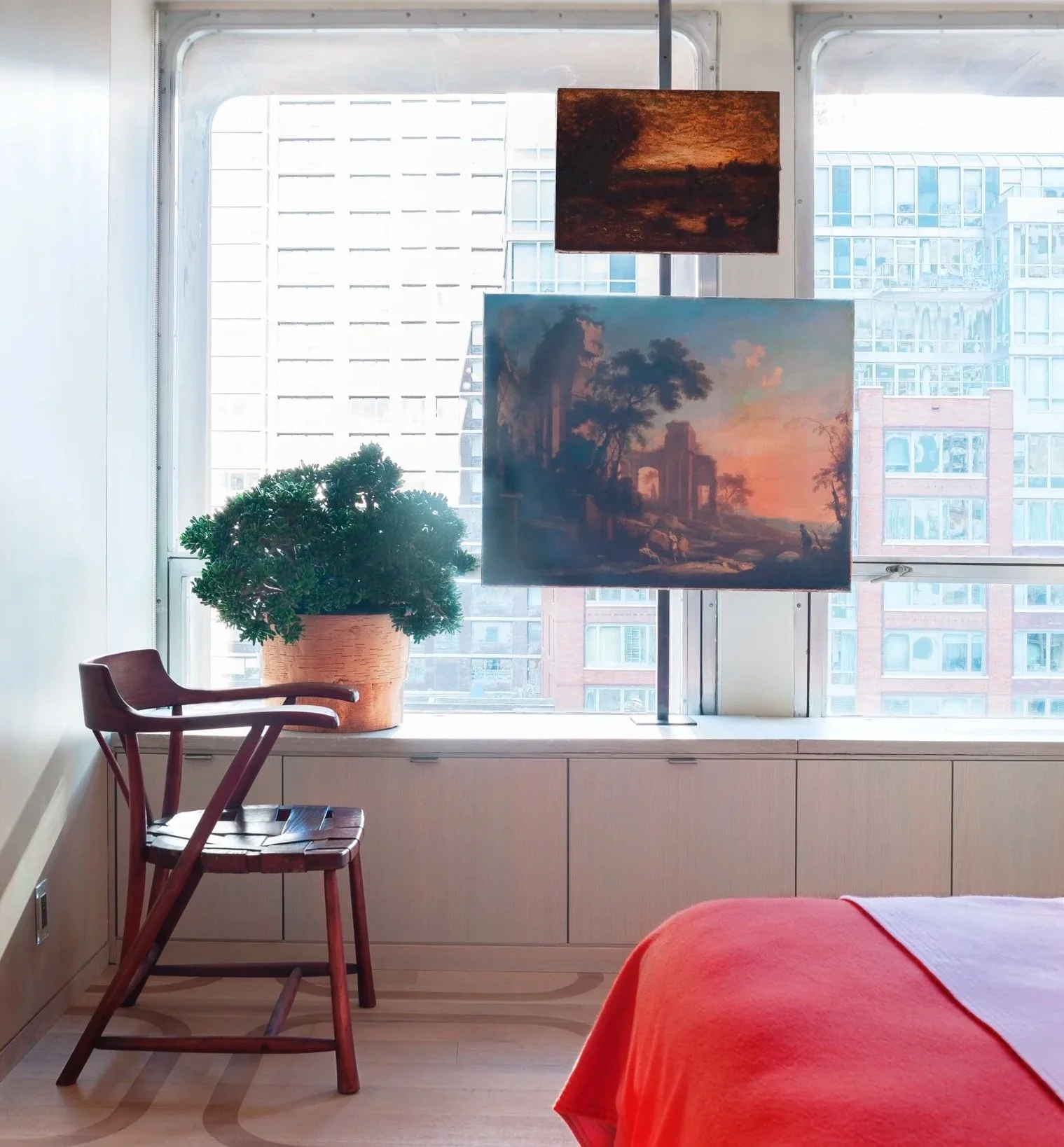 A corner of a room with a large window overlooking a cityscape of tall buildings. There is a wooden chair with a backrest, a potted plant on the windowsill, and two paintings hanging in front of the window. One painting depicts a sunset or sunrise sc