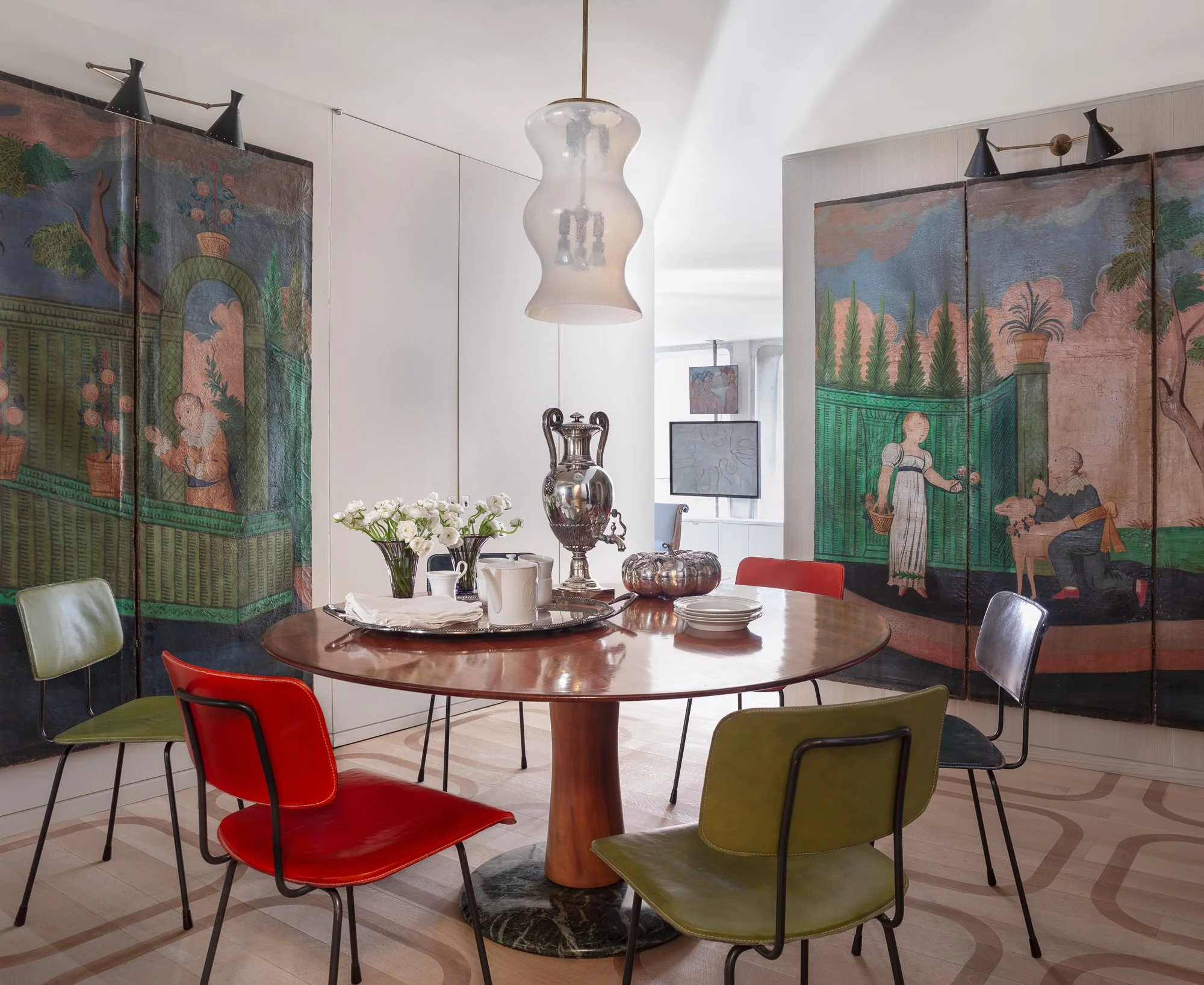A dining area with a wooden round table, colorful chairs, a flower arrangement, silver tea set, and decorative art panels with a garden scene on the wall.