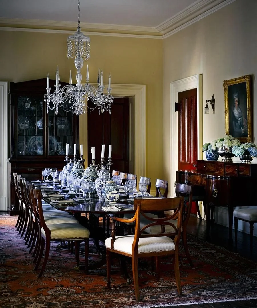 Elegant dining room with a long wooden table set with blue and white porcelain dishes, crystal glasses, candles, and a crystal chandelier overhead. There are wooden and upholstered chairs around the table, a wooden china cabinet, and a sideboard with