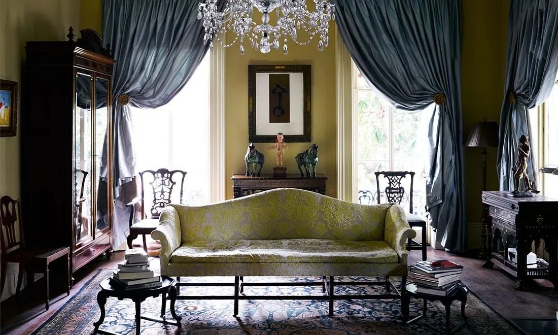Elegant living room with green walls, large windows with dark blue curtains, a chandelier, a vintage sofa, various antique furniture, and decorative art and sculptures.