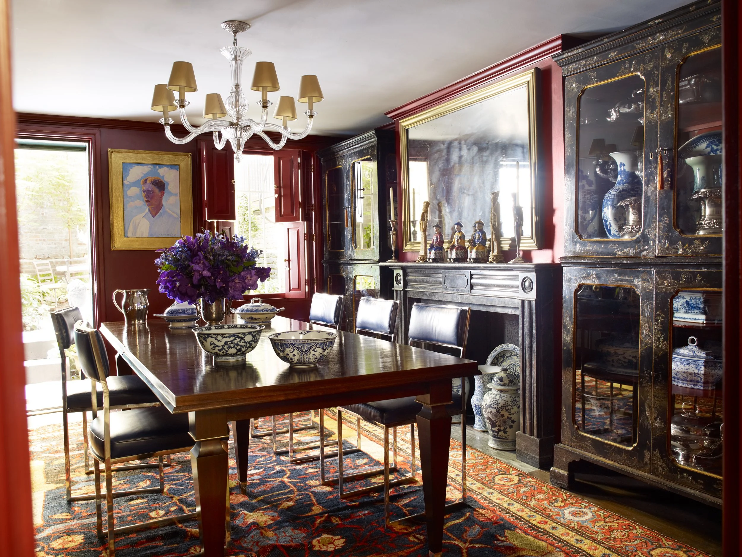 Dining room with a wooden table, black chairs, blue and white china, a vase of purple flowers, a large mirror, black and gold cabinets, and a chandelier, with red walls and a rug.