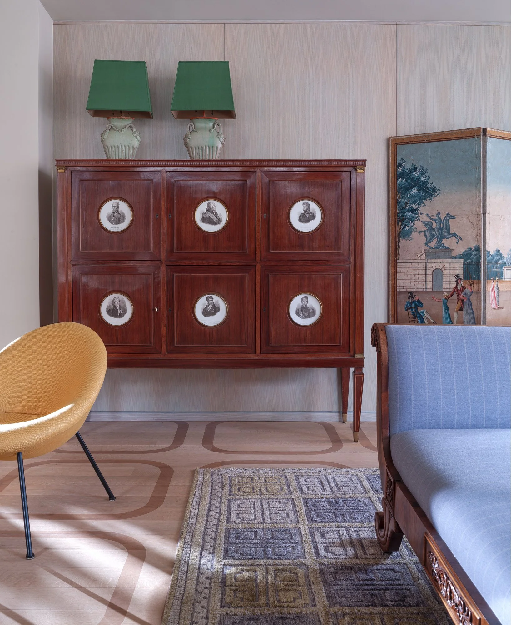 Living room with a wooden cabinet decorated with six black-and-white portrait photos, two green lamps on top of the cabinet, a yellow chair, part of a blue sofa, a patterned rug, and a decorative folding screen with a scenic painting.
