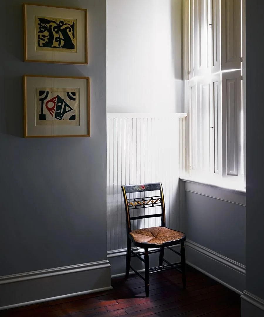 Sunlit corner of a room with a wooden chair with woven seat, white walls, colorful abstract artwork on the wall, and an open window with white shutters letting in sunlight.
