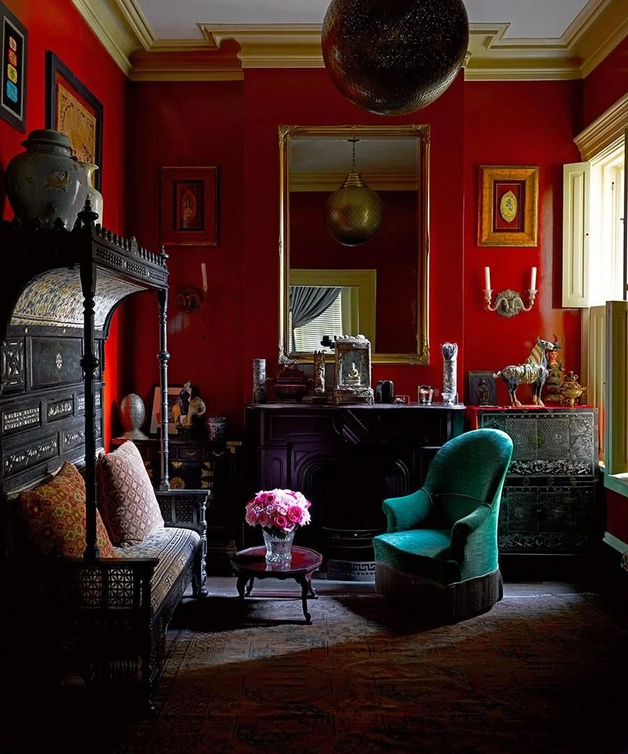 A richly decorated room with red walls, gold accents, and vintage furniture. There is a green upholstered armchair, a small table with pink flowers, a dark wooden cabinet with decorative items, and various framed artworks. A large mirror is on the wa