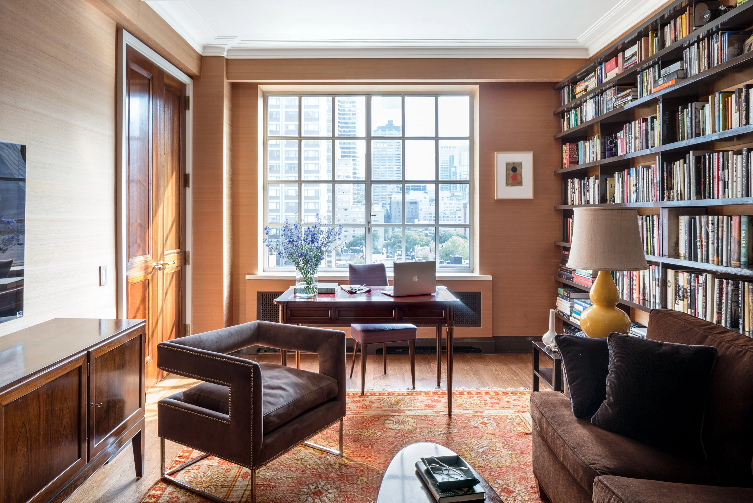 A well-lit living room with a large window overlooking a cityscape, a wooden desk with a laptop and flowers, a brown leather chair, a beige couch with pillows, a yellow lamp, a bookshelf filled with books, and a patterned rug.