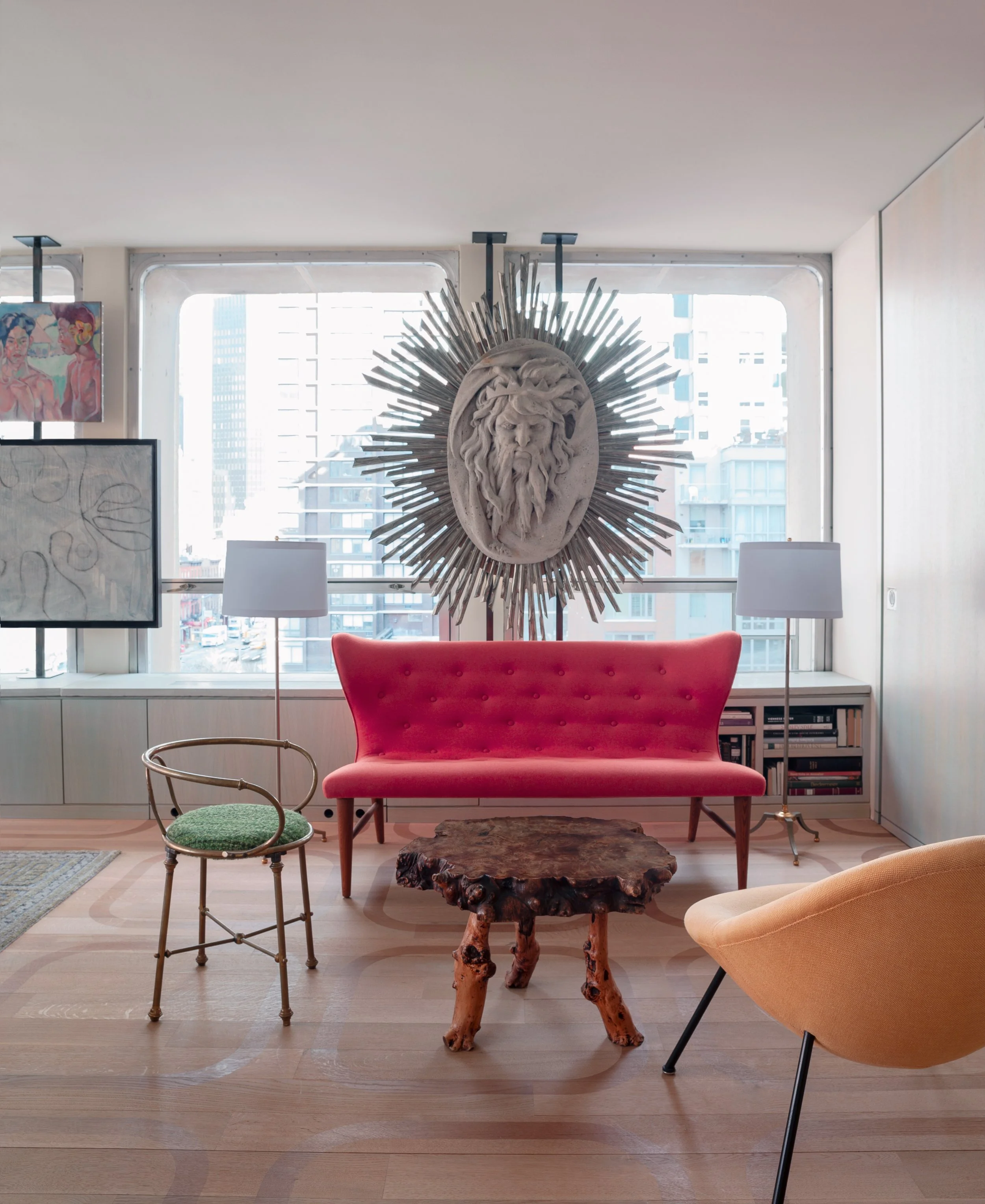 Interior of a modern living room with large windows, a large lion head mural with sunburst design, pink velvet sofa, wooden coffee table, two floor lamps, and various artworks.