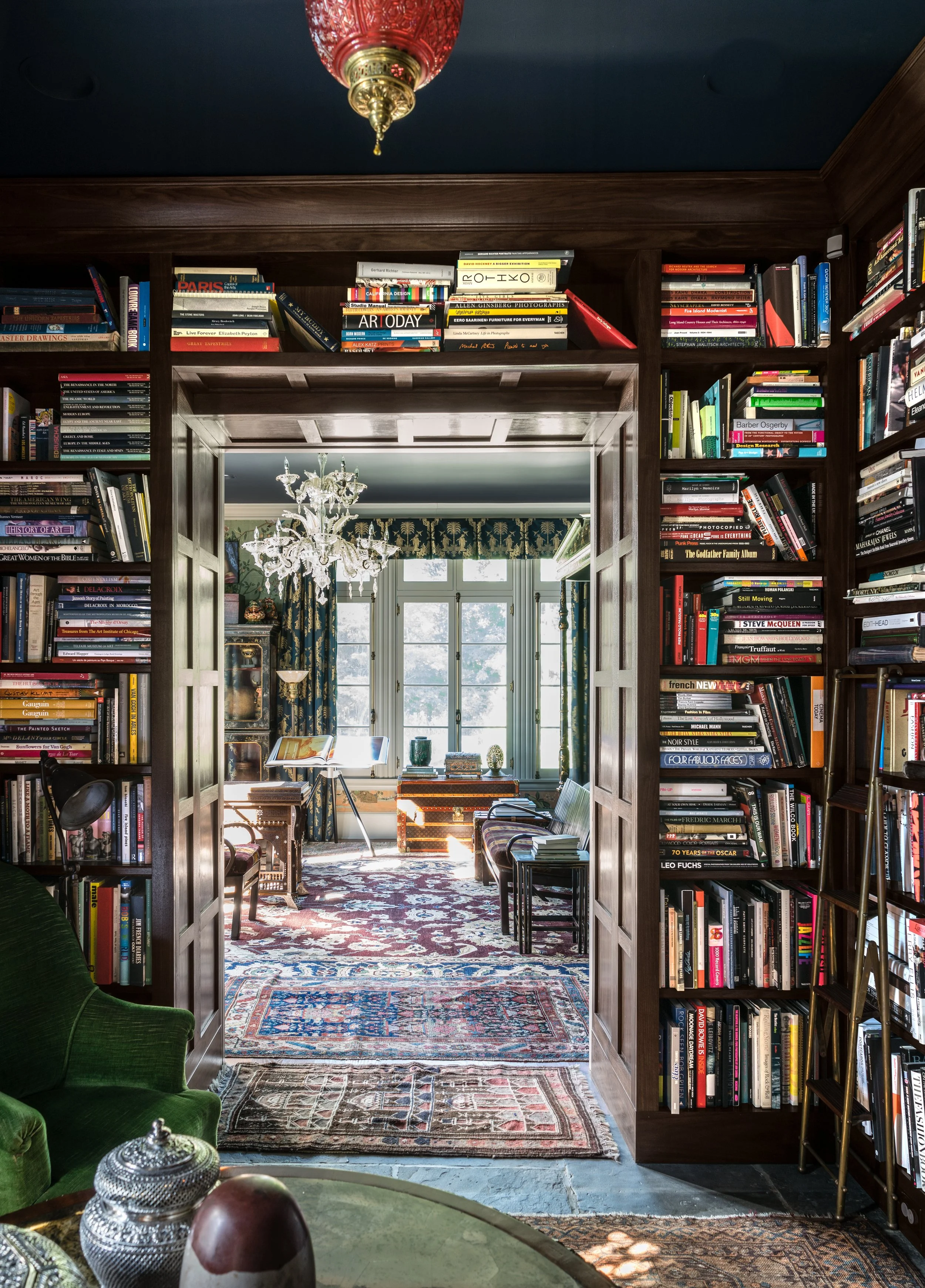 View through a doorway into a bright, elegant room with large windows, a chandelier, and Victorian-style furniture, surrounded by bookshelves filled with books.