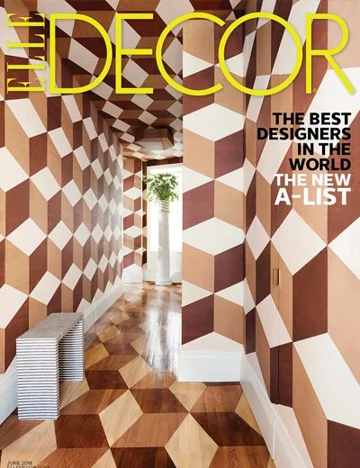 Interior hallway with geometric-patterned walls and floor, featuring brown and white shapes, a white stand with a green plant, and stacked white books, in a modern decor style.
