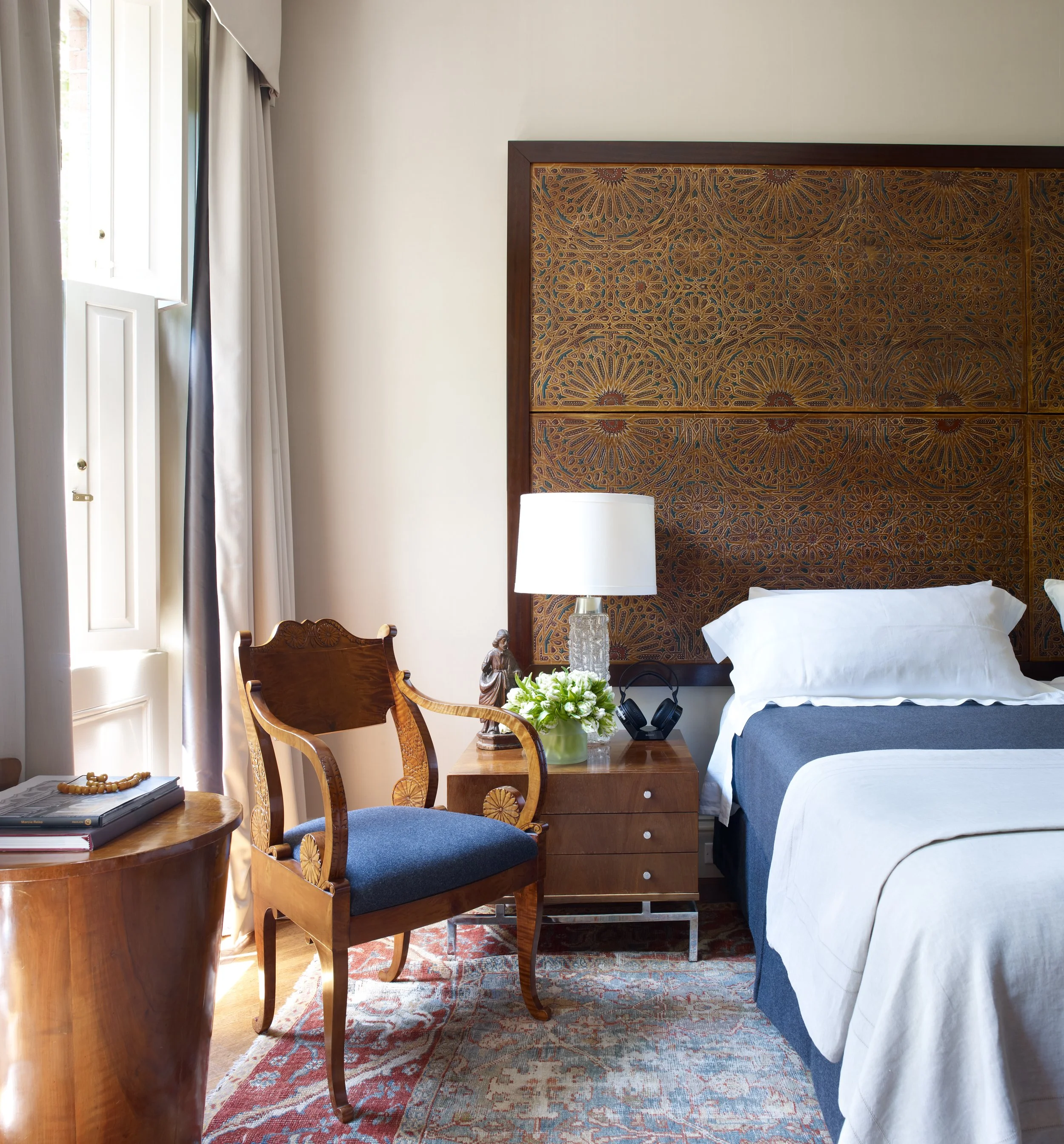 Hotel room with a large headboard, bedside table with lamp, flowers, and headphones, chair, and window with curtains.