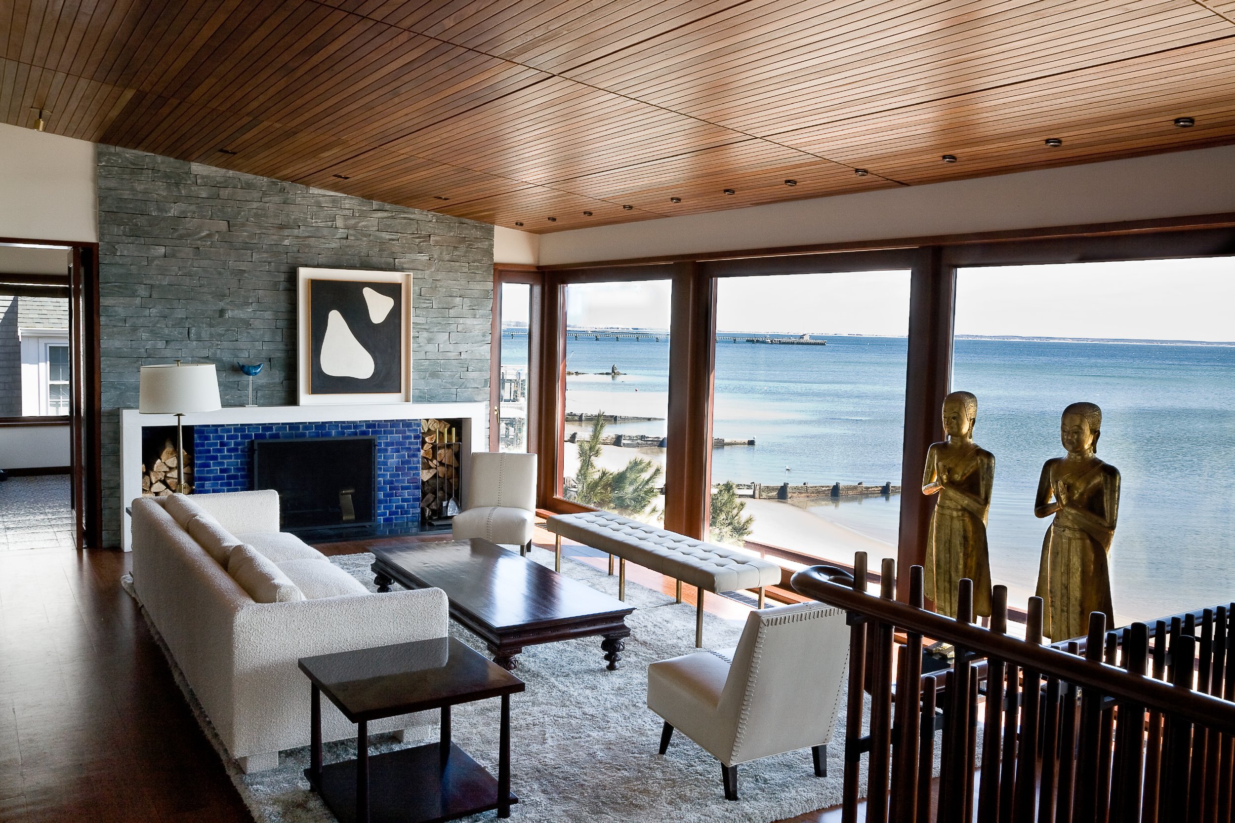 Living room with large windows overlooking the beach and ocean, contemporary furniture, a stone fireplace, and decorative sculptures near the window.