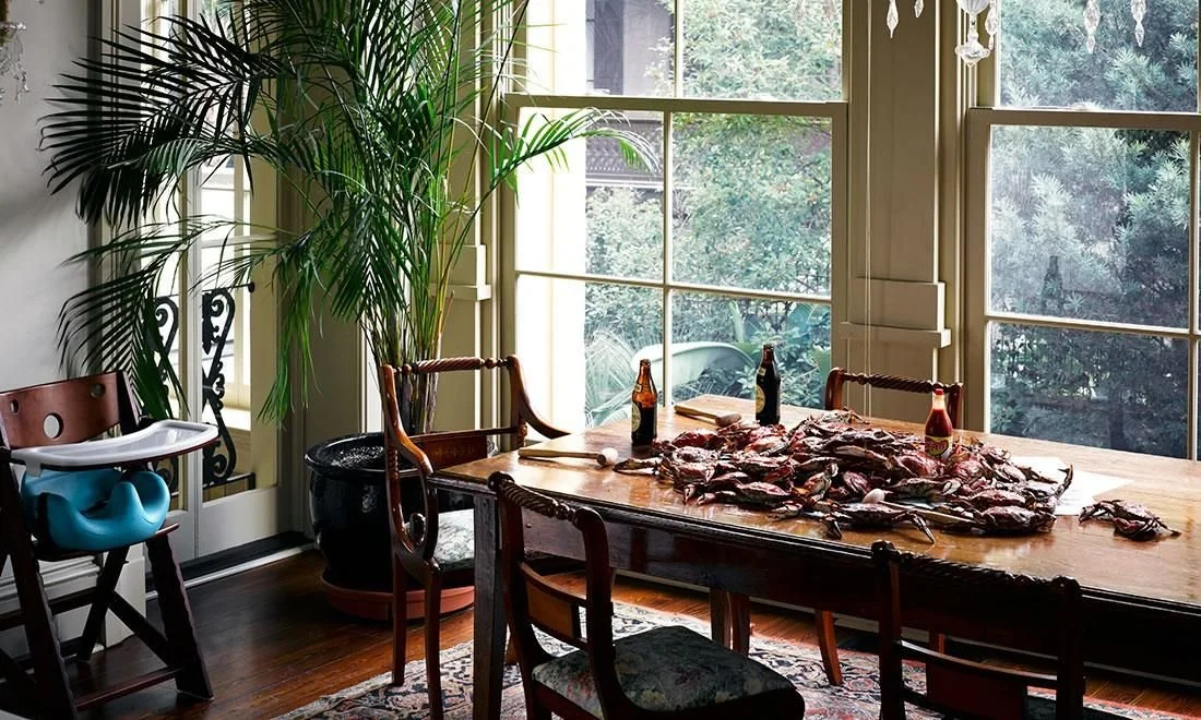 Dining room with a large wooden table covered with raw meat and three bottles, surrounded by chairs, large windows, and a potted plant.