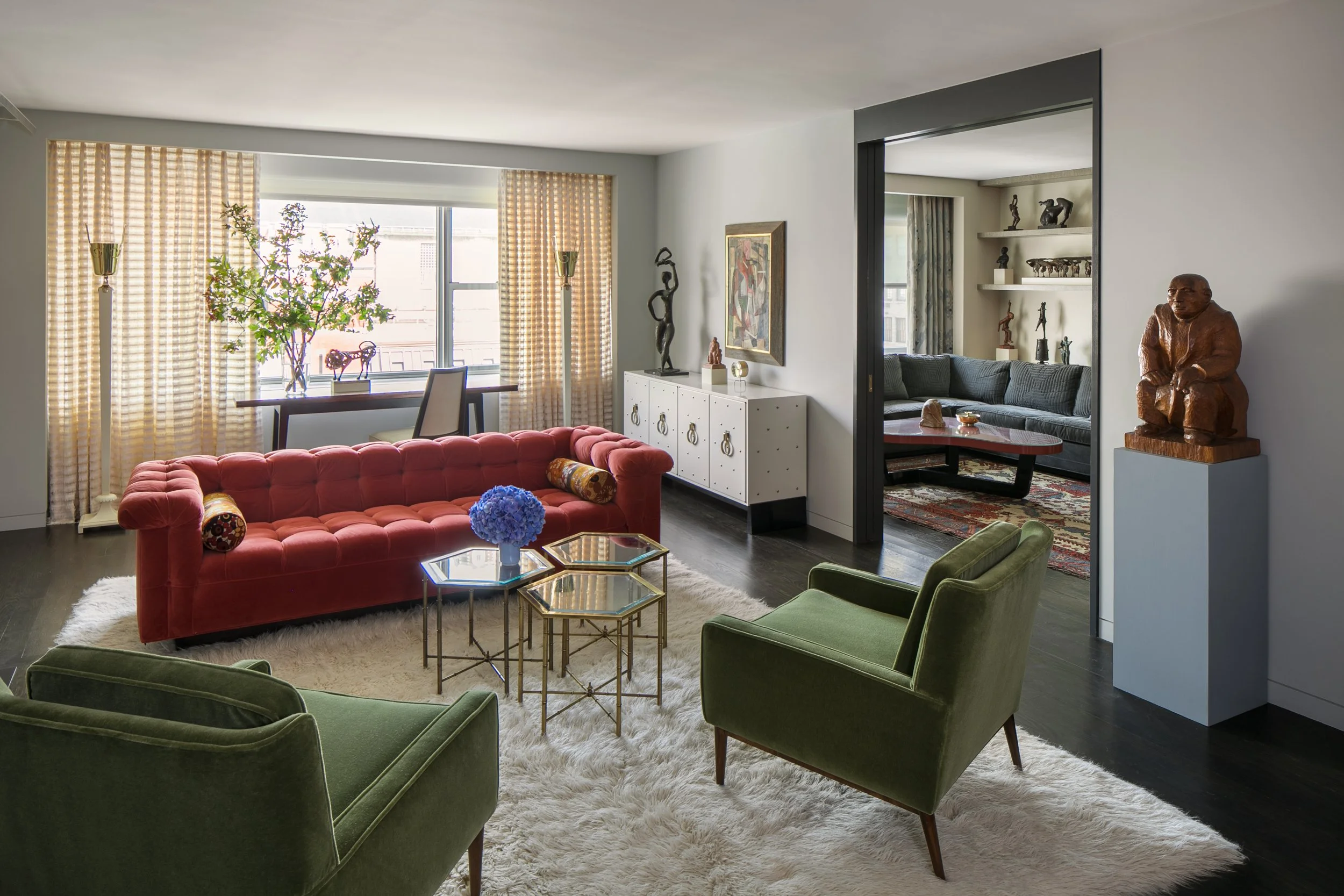 Living room with a red sofa, two green armchairs, glass and gold coffee tables, a white shag rug, a window with yellow curtains, decorative sculptures, artwork, and a doorway leading to another seating area.