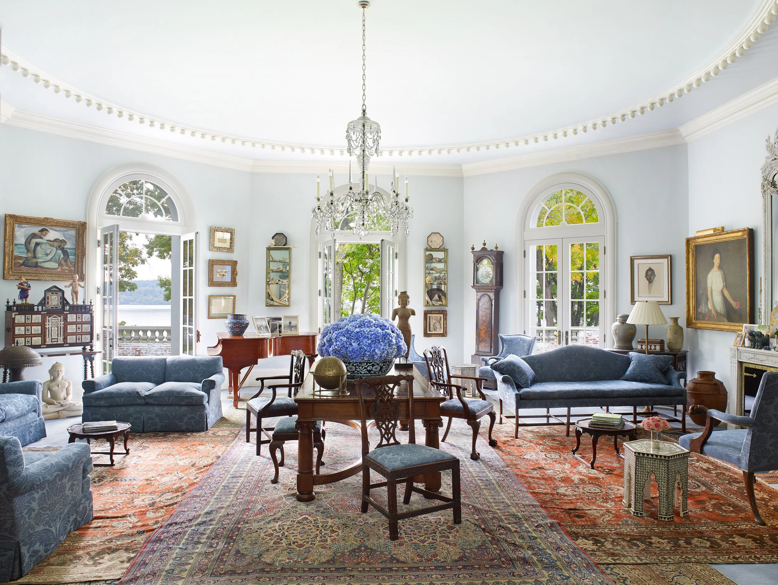 Elegant living room with blue sofas, antique furniture, framed artwork, a grand chandelier, and large arched windows showing greenery outside.