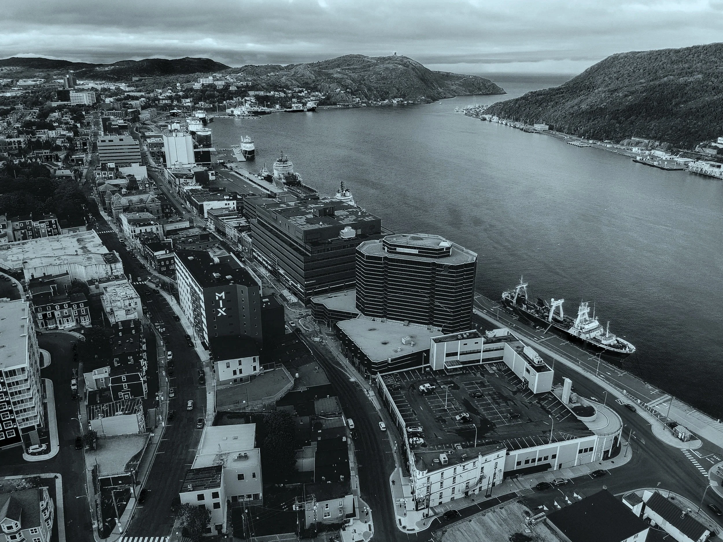 Aerial view of a Newfoundland harbour with boats docked along the waterfront, surrounded by urban buildings and hilly terrain in the background.