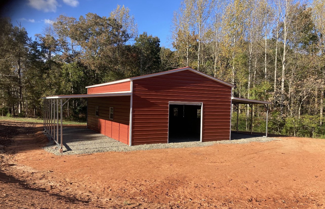 Red metal barn with open garage doors, surrounded by trees and a dirt lot.