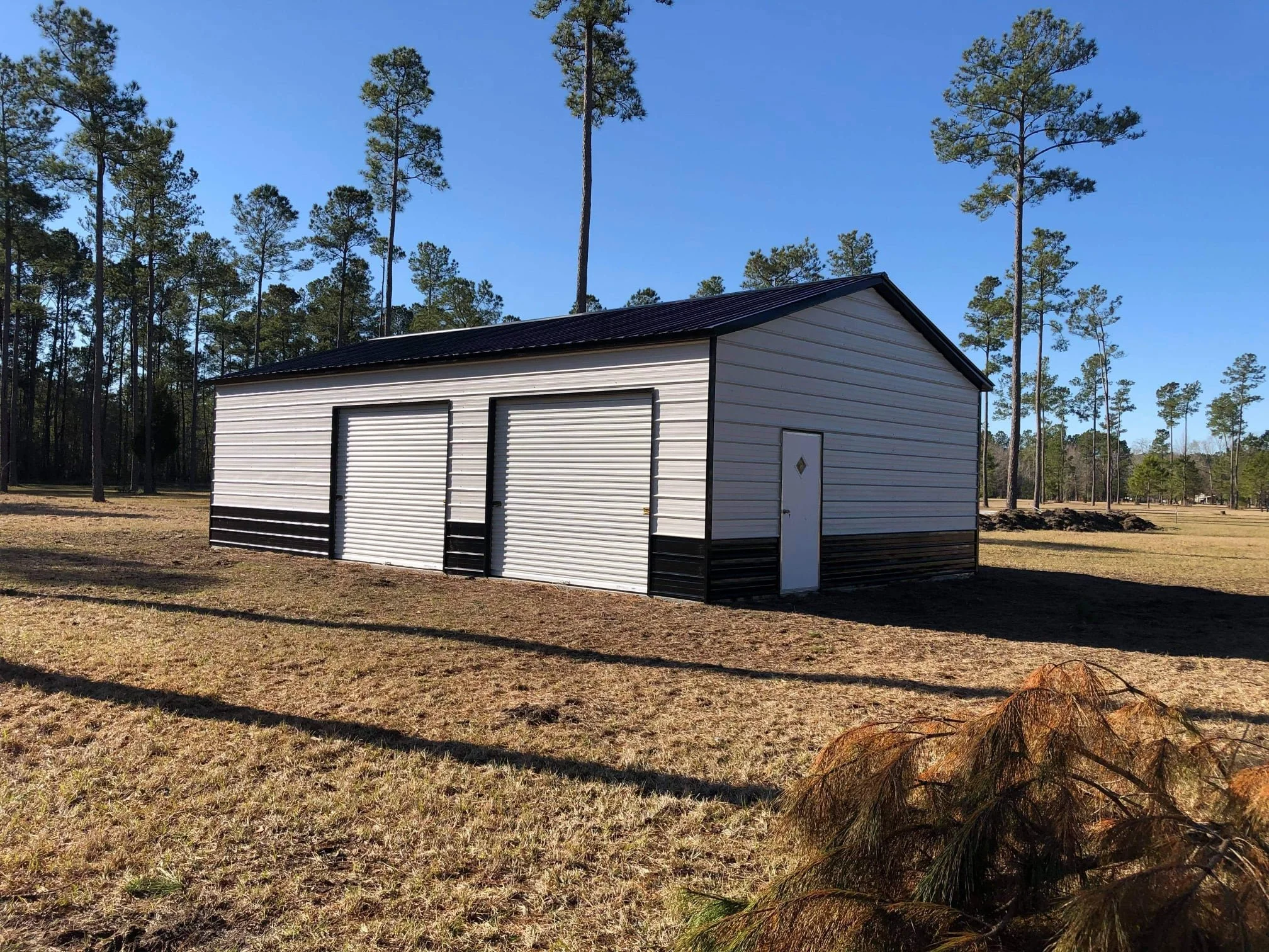 Large outdoor metal shed with two roll-up garage doors and a single side door in a grassy area surrounded by tall pine trees.