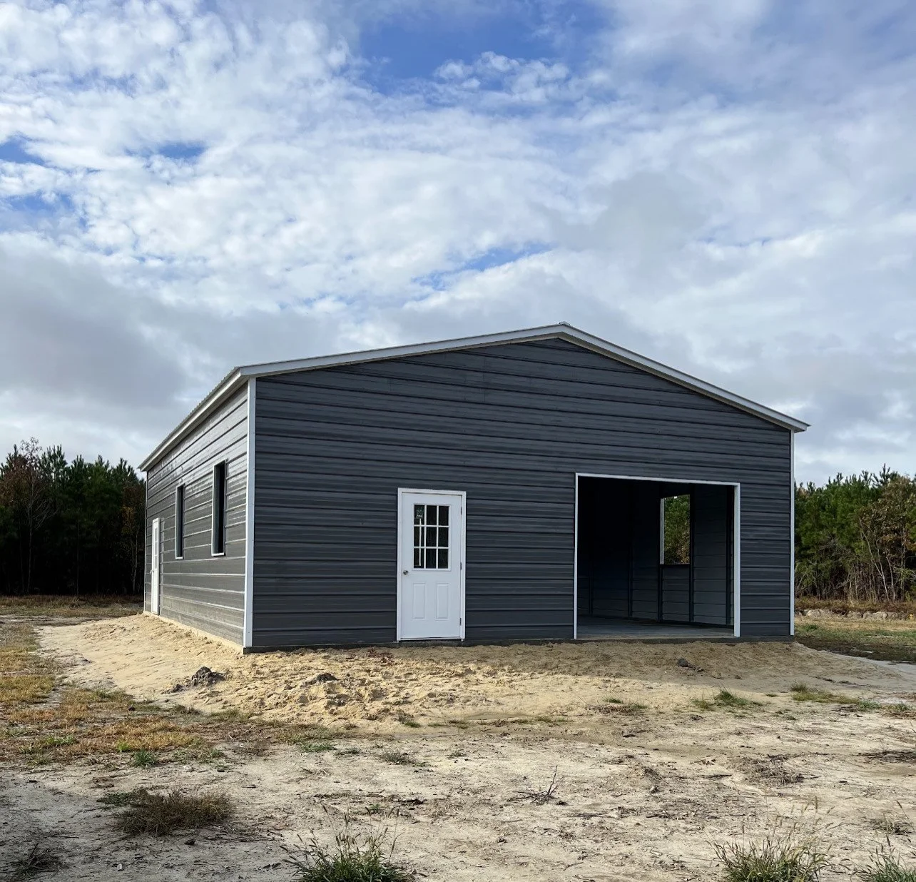 Gray metal building with a white door and open garage-style space, set on sandy ground with trees in the background under a partly cloudy sky.