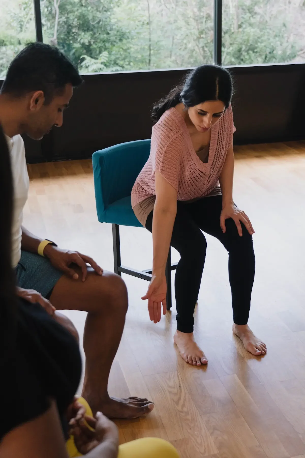 Geeti, a trauma-informed yoga therapist, demonstrating a seated stretch or exercise to a group of people in a room with large windows and wooden floors.