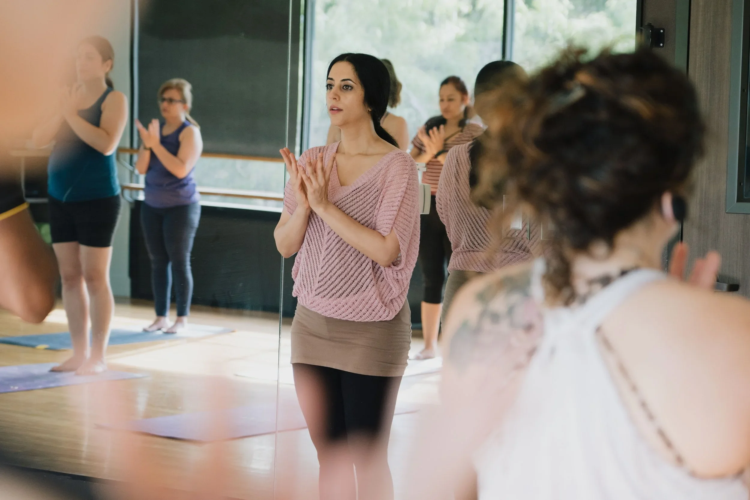 Women participating in trauma-informed yoga or meditation class, standing on mats and following instructions, with a studio featuring large windows and a mirror.
