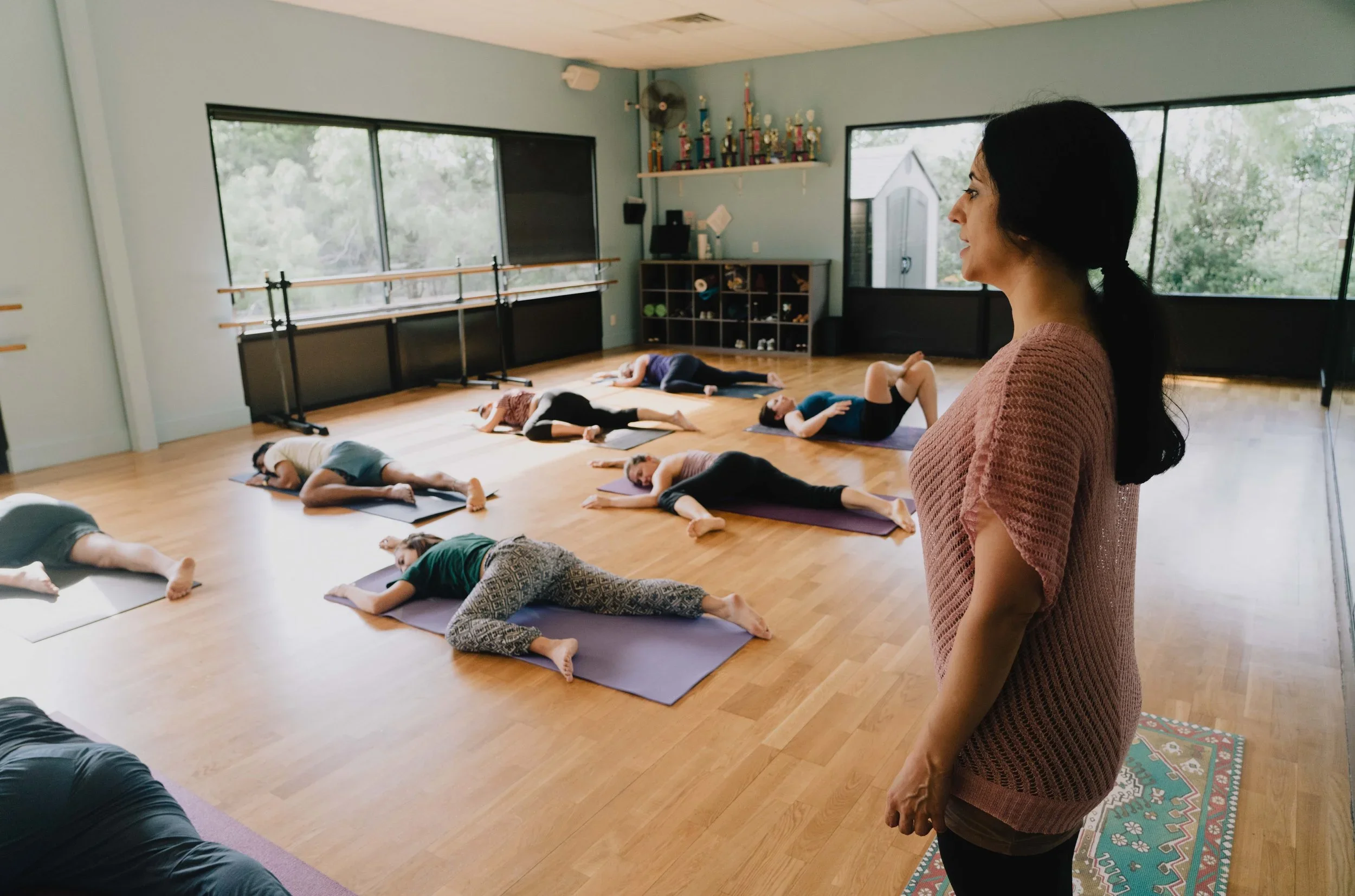 A yoga instructor standing in a studio observing students lying on mats practicing yoga.