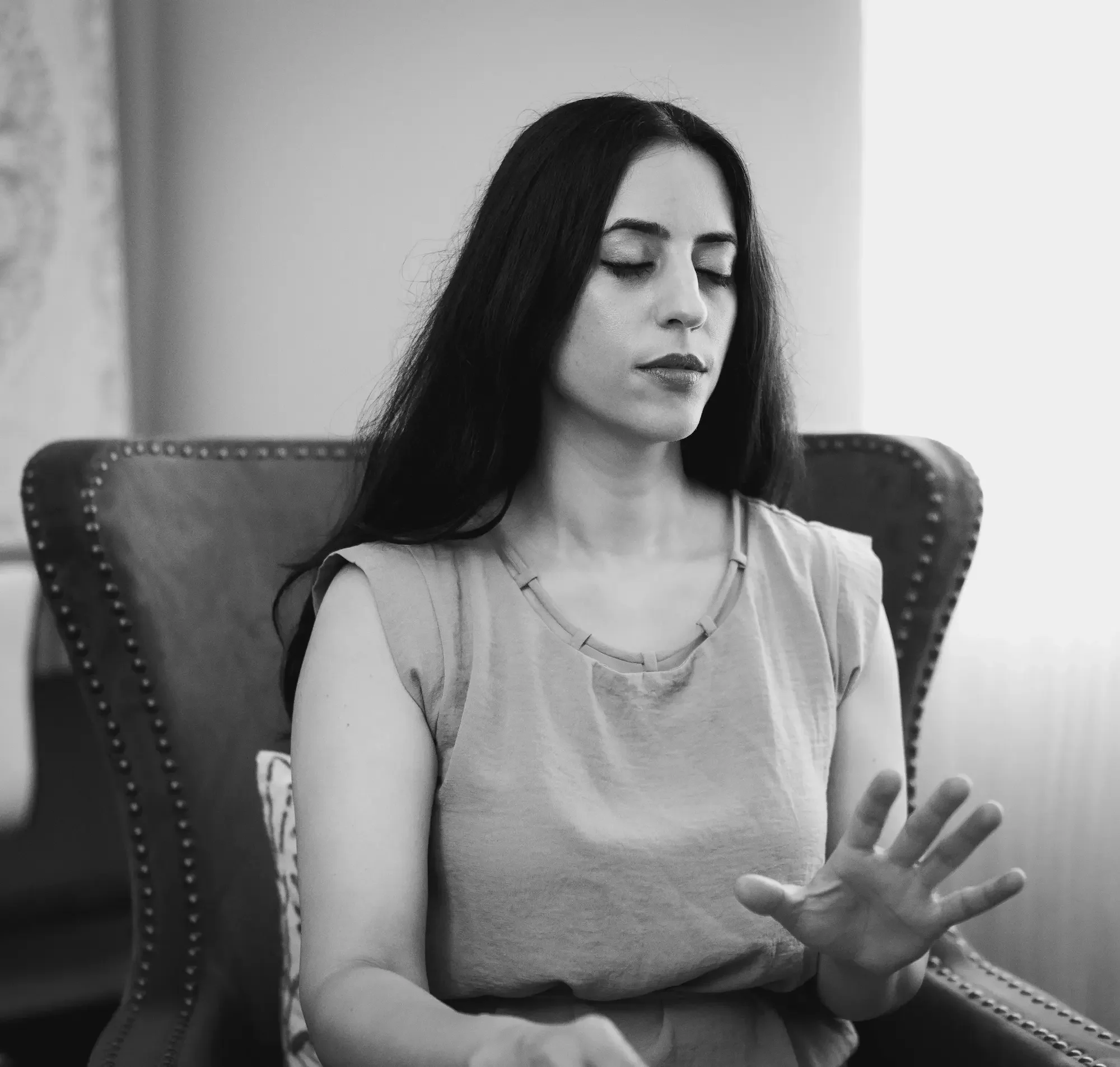 An energy healer with long dark hair sitting in an armchair with her eyes closed, holding her right hand up with her palm facing outward.