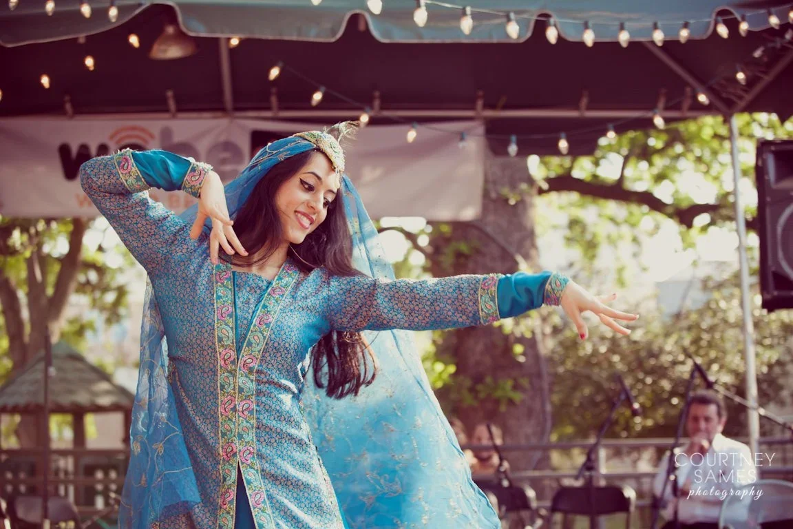 A woman dressed in traditional South Asian attire dancing outdoors on a stage with string lights, trees, and musicians in the background.