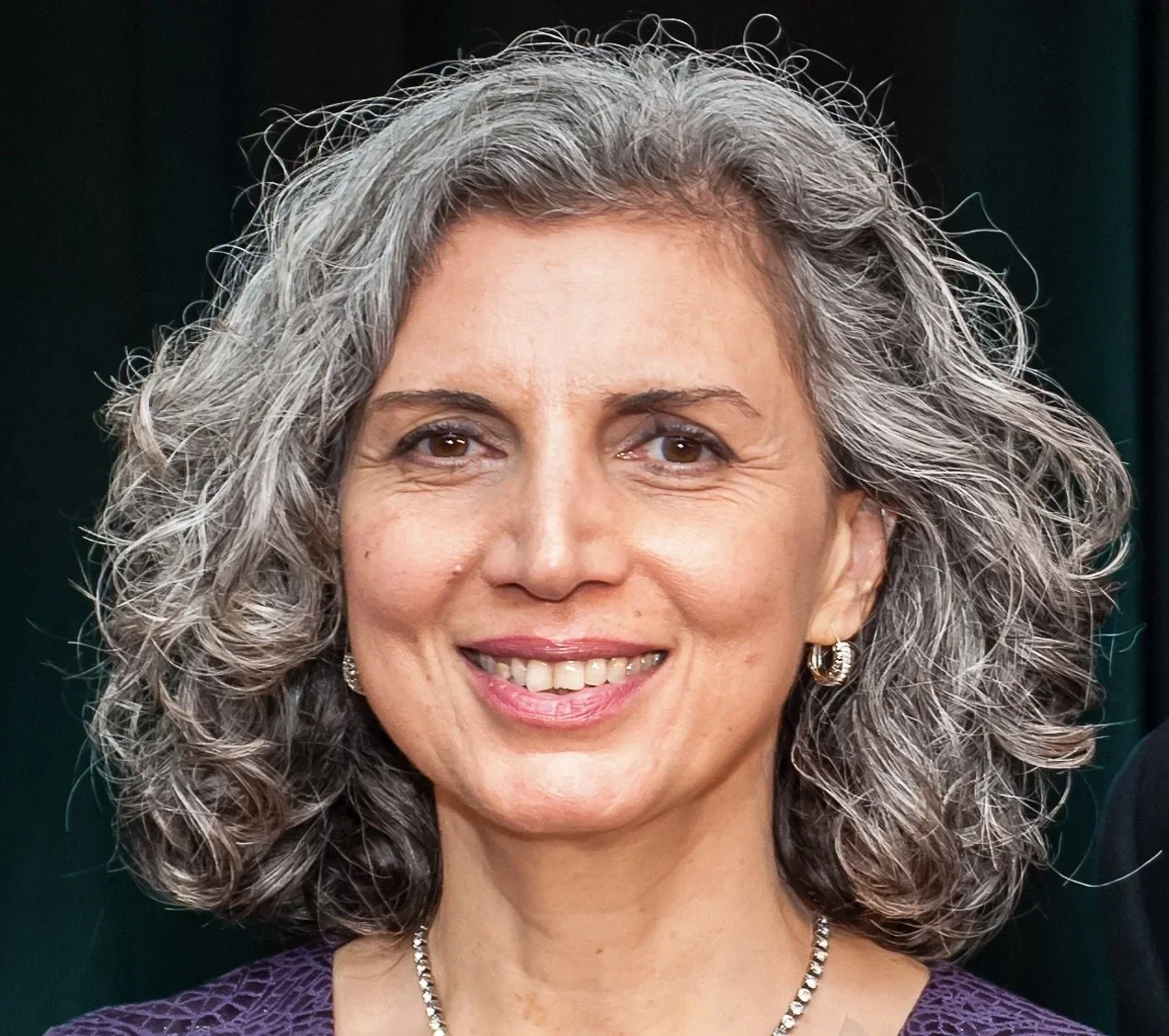 A trauma-informed yoga participant smiling with shoulder-length curly gray hair, wearing earrings, a necklace, and a purple outfit.