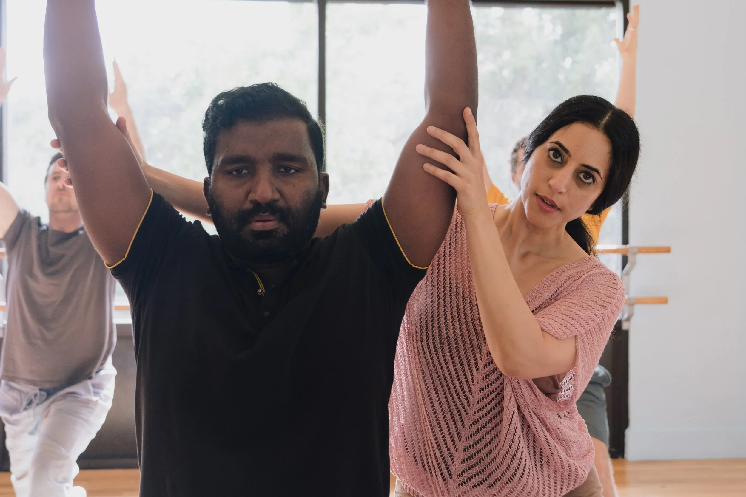 A diverse group of people participating in trauma-informed yoga therapy with Geeti assisting a man with his stretch in a bright studio.