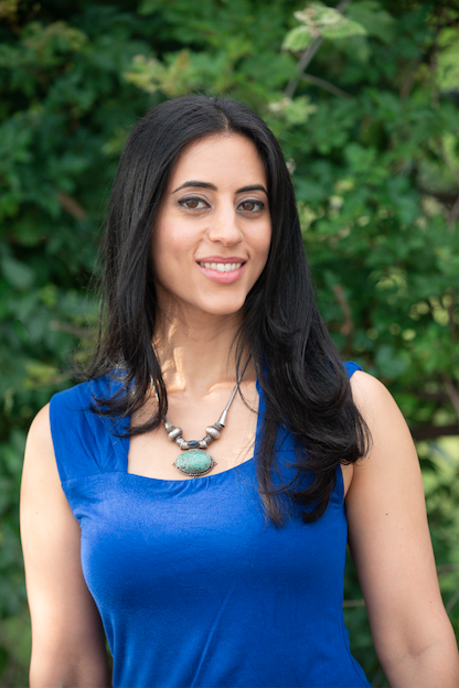 An energy healer with long black hair, wearing a bright blue sleeveless top and a statement necklace with a large turquoise stone, standing outdoors with green foliage in the background.