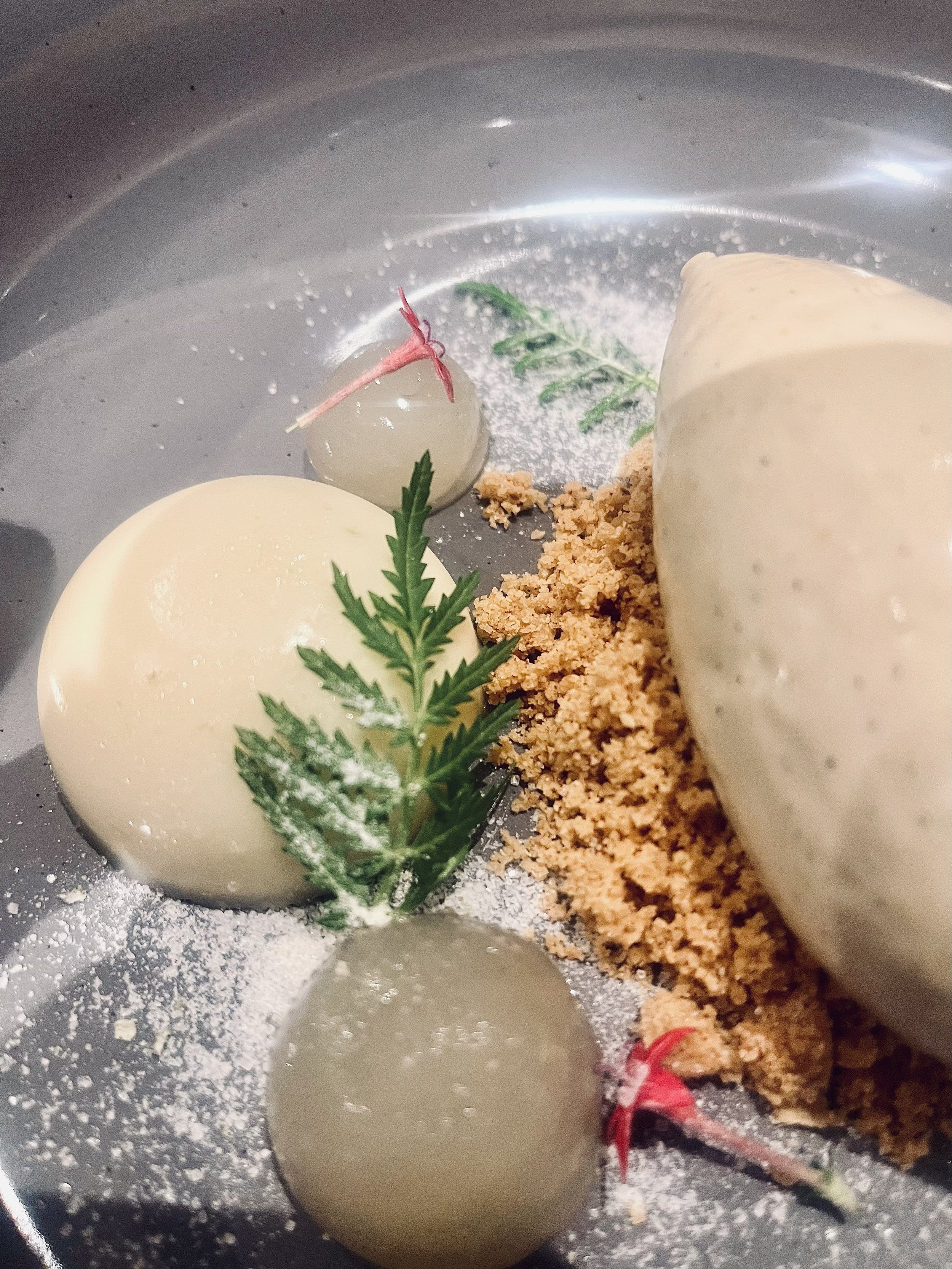 A gourmet dish featuring two spheres of white cheese or mousse, garnished with a green herb, along with small round balls decorated with pink ribbons and sprigs of greenery, served on a metal plate with a crumb-like substance.