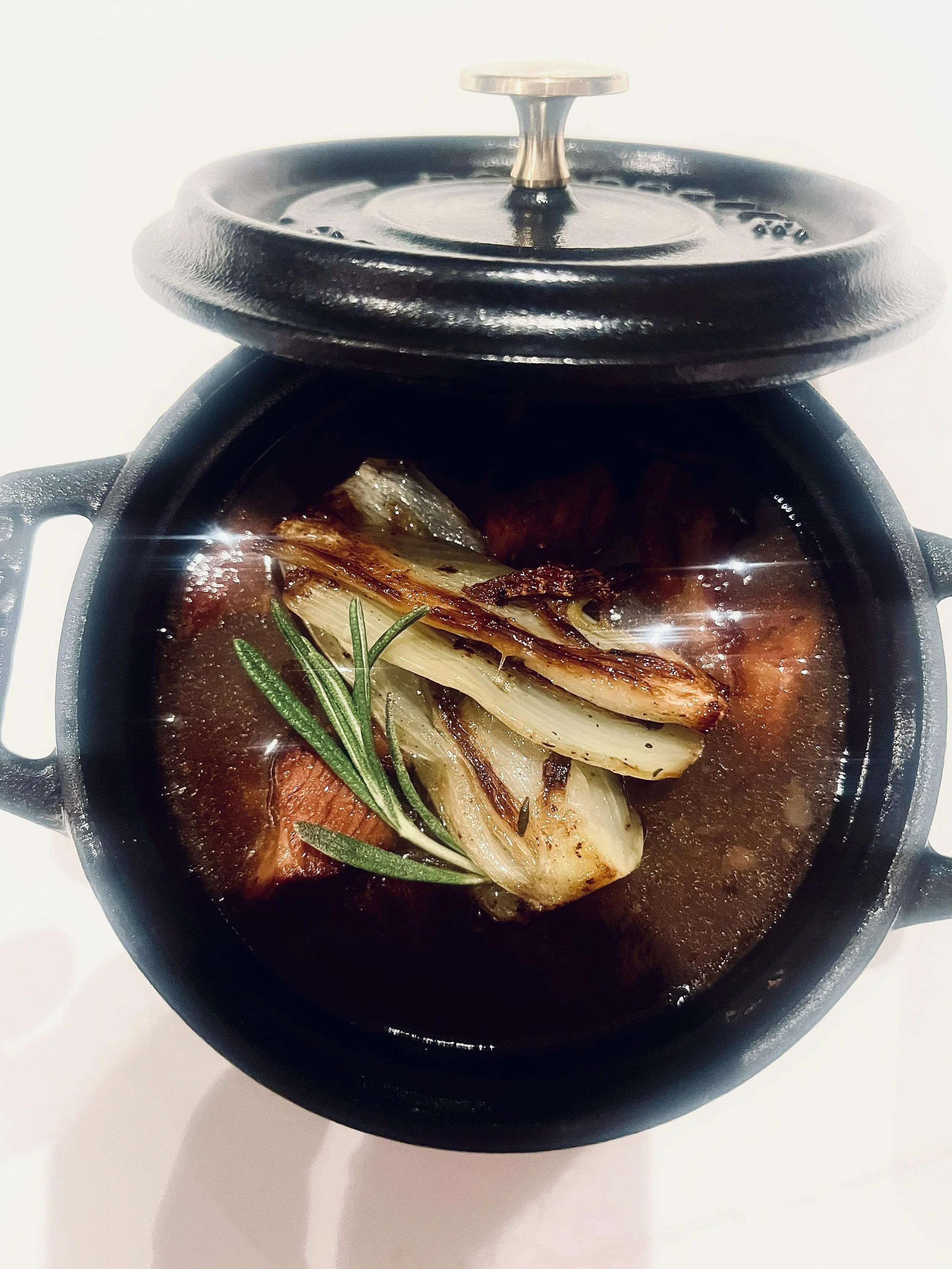 A black cast iron stew pot with a lid slightly open, showing roasted vegetables and herbs in a dark broth.