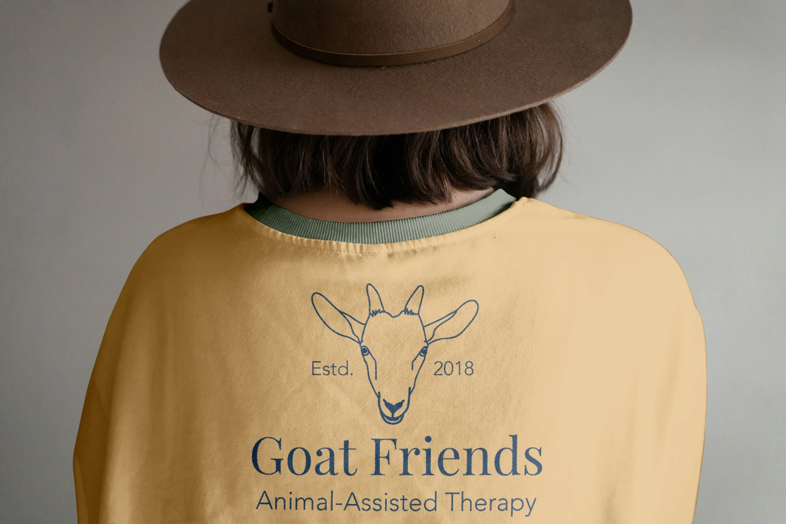 All navy Goat Friends logo is mocked up on back of yellow t-shirt, worn by someone with short brown hair and a brown hat.