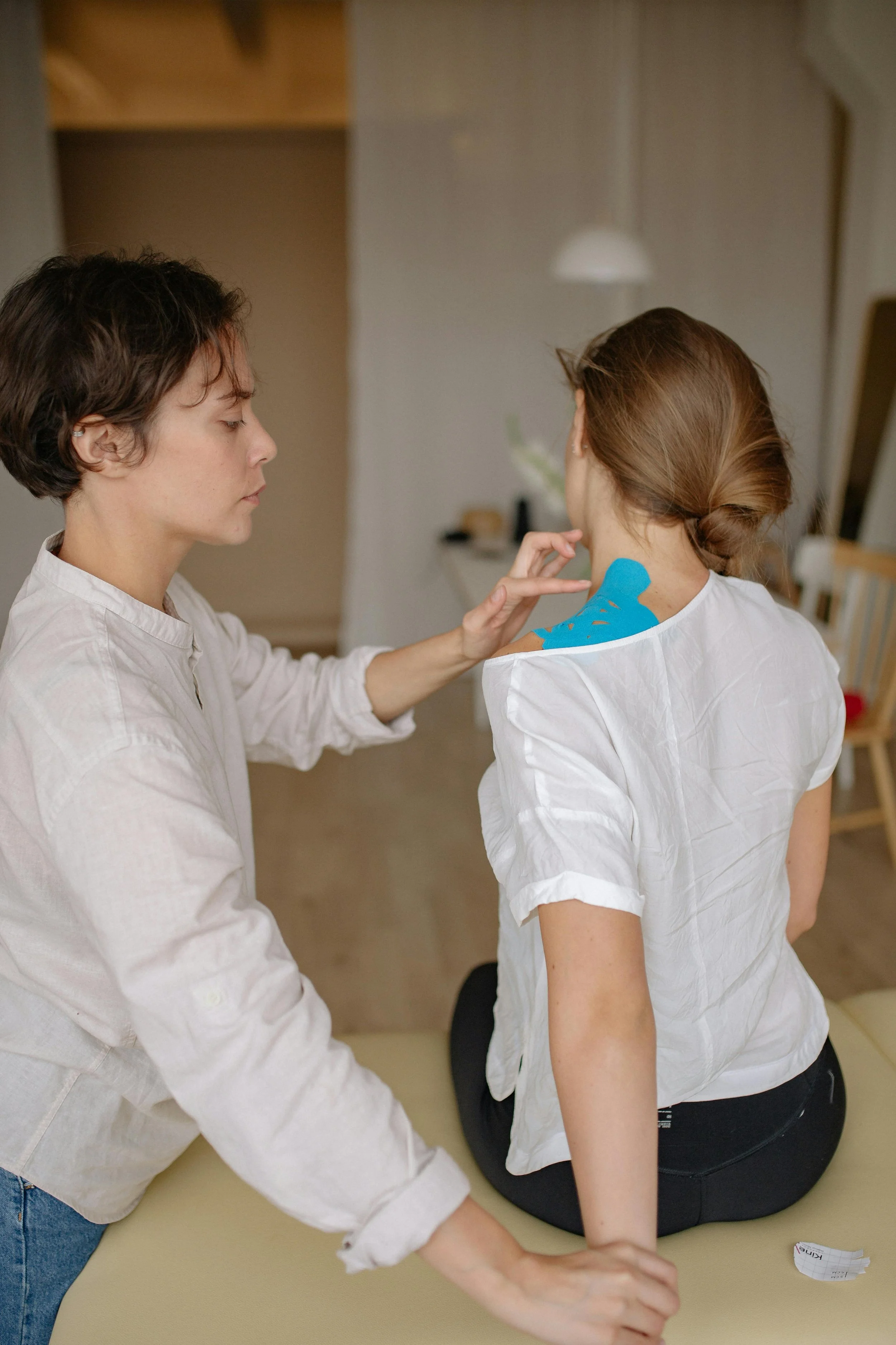 A physical therapist in a white shirt on the left applies KT tape to a patient's neck and shoulder area. Patient has back turned to camera and is wearing a white shirt.