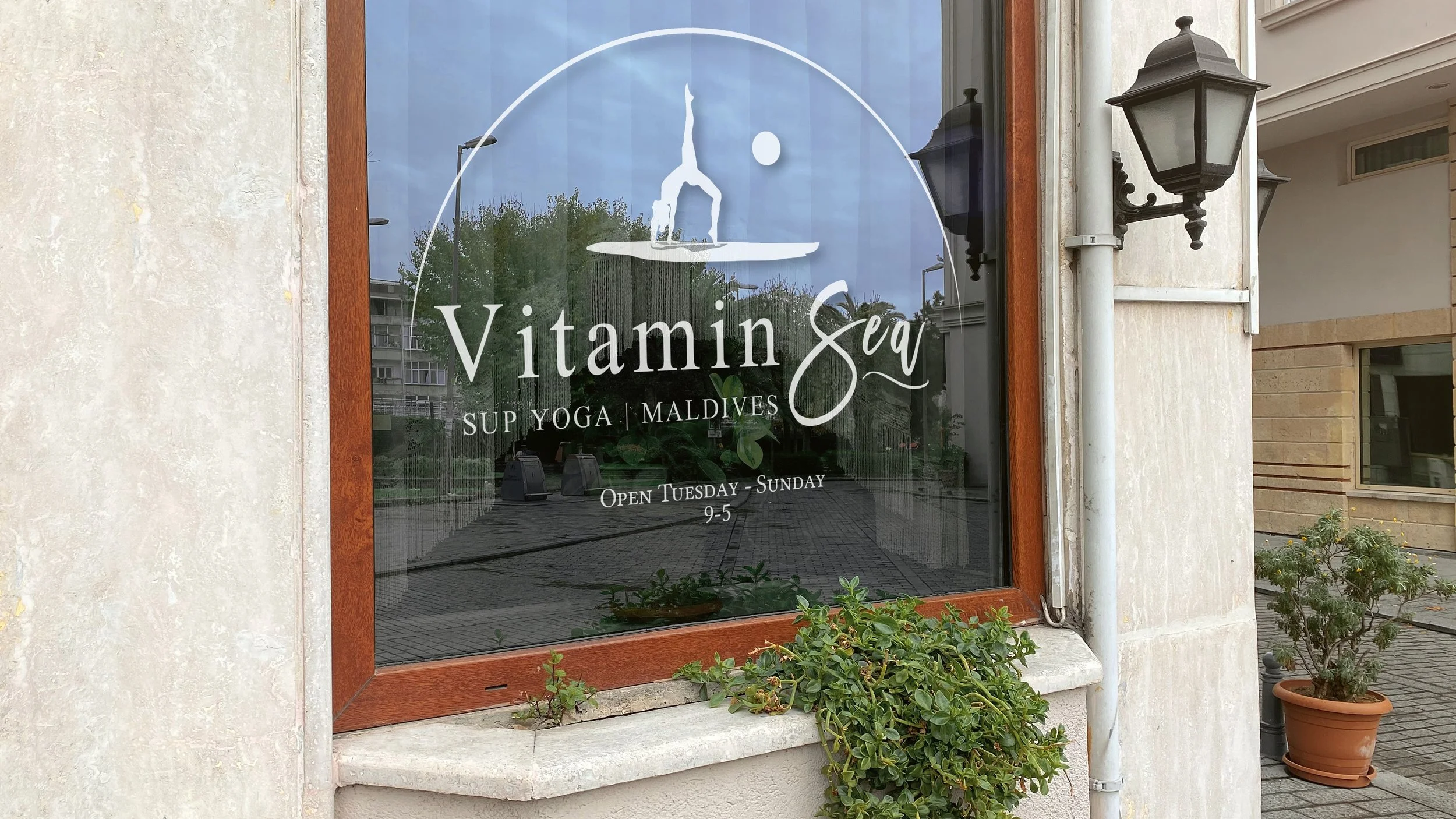 On a glass window, all white business logo is shown as vinyl sticker. Logo: Paddleboarder in yoga pose with sun to right and semi circle above. Business name below: "Vitamin Sea SUP YOGA | MALDIVES" with business hours below.