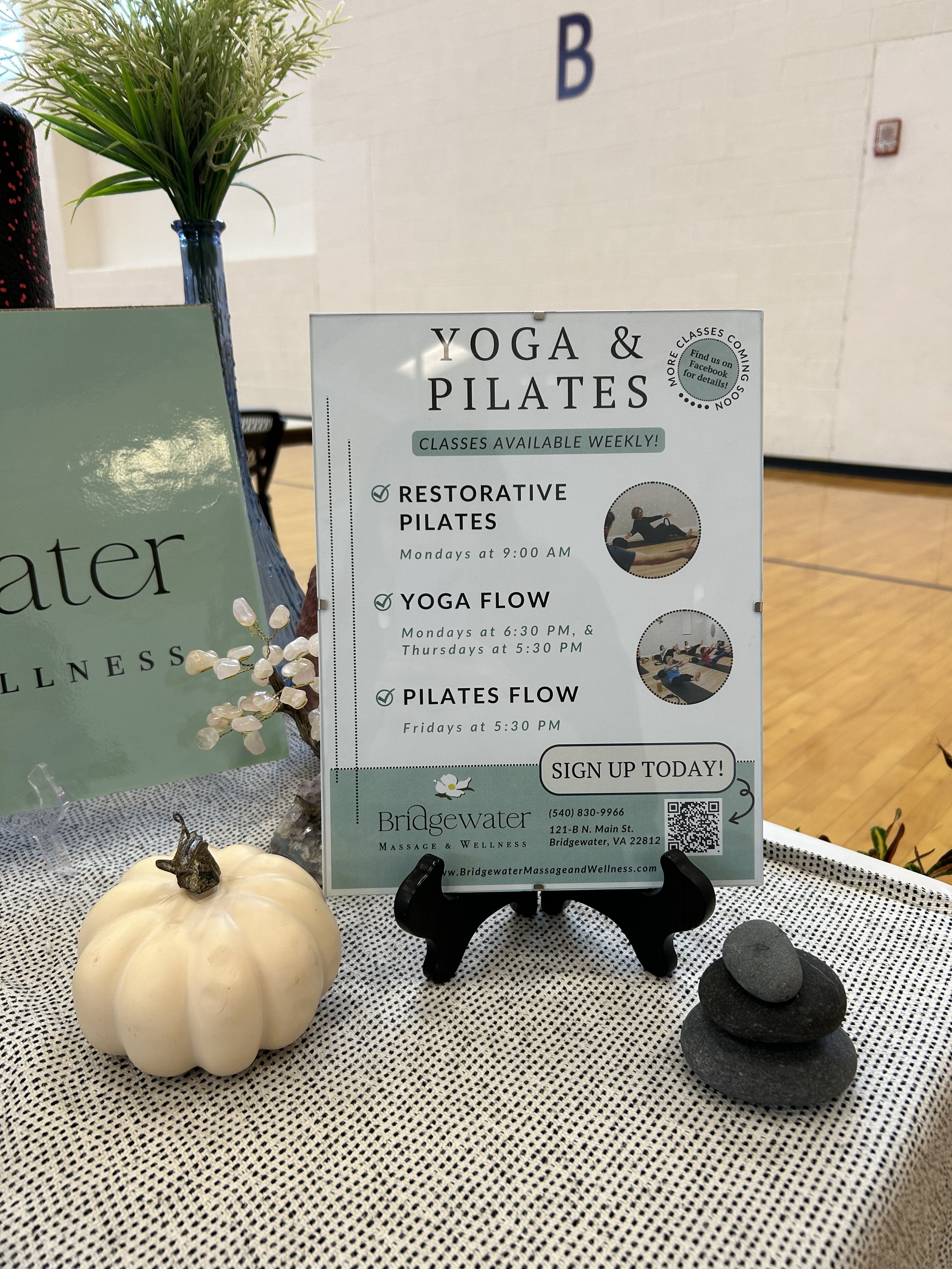 A framed sign reads "YOGA & PILATES" with the class schedule, images from class, and additional marketing information. Sign is in a stand on table with gray and white cloth, white pumpkin decor, and background table decor.